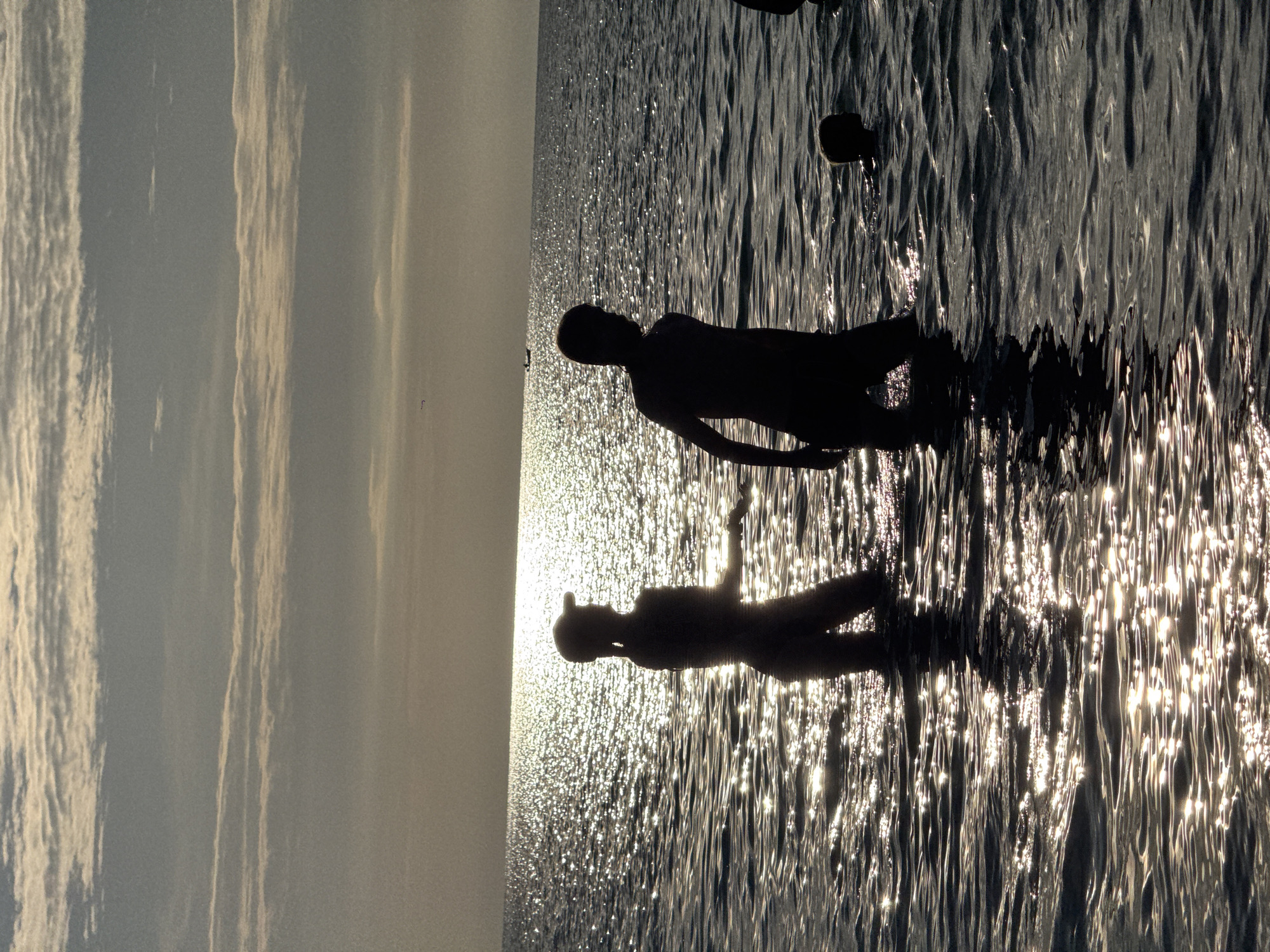 Two silhouettes of people are standing in the ocean with sunlight glinting off the water. On the left, a person wearing a cap stands facing to the right. On the right, a person stands facing left. In the background, a small boat is visible on the horizon under a sky with scattered clouds. The scene is set at the beach in ตำบลเกาะพะงัน, Thailand.