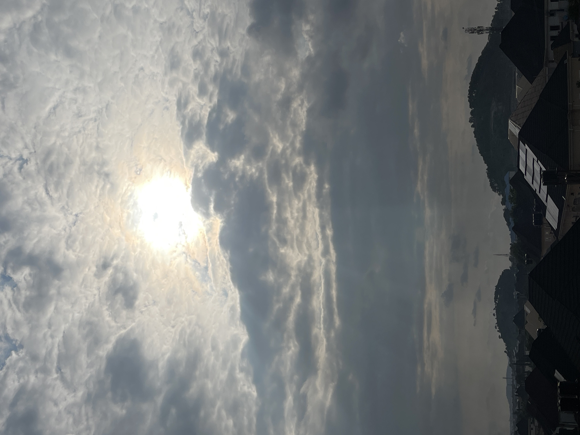 The image captures a dramatic sky over the cityscape of Abuja, Nigeria. The sun is partially obscured by a thick layer of textured, grey clouds, creating a diffused, almost ethereal glow. Hints of blue sky can be seen through thinner patches of clouds higher up. Below the sky, the distinct silhouettes of rooftops of buildings, mostly dark and angular, dominate the foreground and mid-ground, suggesting a residential area. Further in the distance, rolling hills, covered in sparse vegetation, rise above the urban sprawl. A prominent hill with a rounded summit, dotted with what appear to be trees or structures, is visible in the center-right of the frame. A cellular tower stands tall on the right side of this hill. The overall atmosphere is moody and overcast, indicative of a cloudy day in the city.
