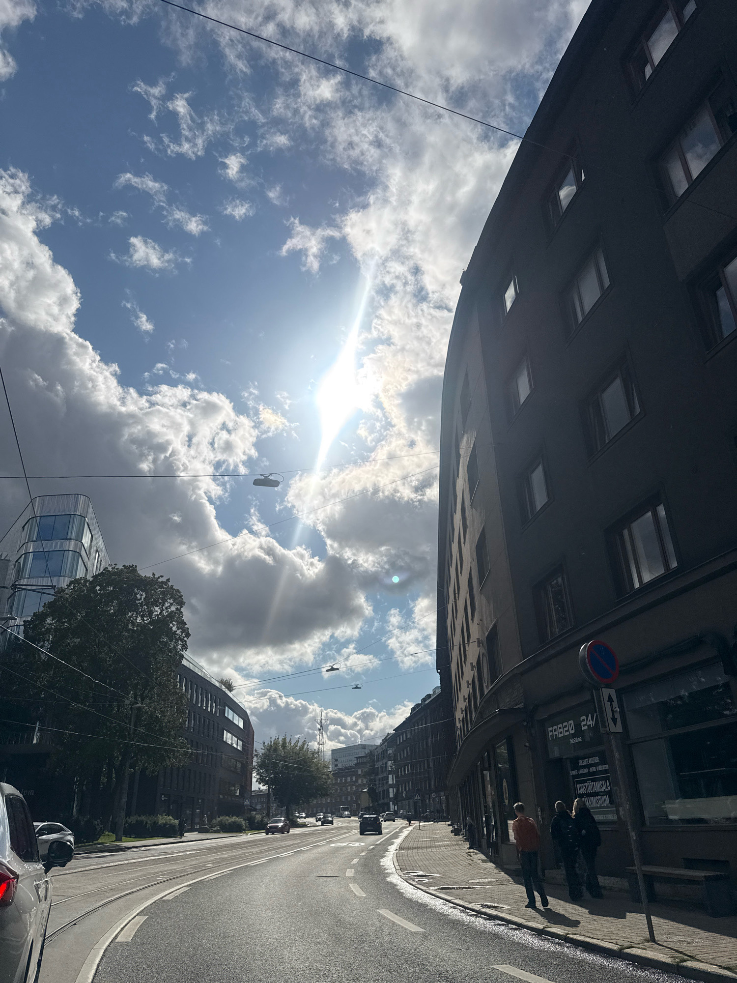 The image depicts a street scene in Tallinn, Estonia, on a bright, cloudy day. The sun is visible, casting a strong glare and creating lens flare in the upper central part of the image. Power lines crisscross the sky. On the right side of the street is a dark, multi-story building with numerous windows. A sign on the building reads "FB20 24/". In front of the building, three people are walking on the sidewalk. A traffic sign with a blue circle and red diagonal line is mounted on a pole near the building. The street in the foreground curves to the left and has tram tracks. Cars are present on the road and a silver car is parked on the left. To the left of the street, there are trees and other buildings, including a modern building with a glass facade. The ground is wet in some areas, suggesting recent rain.