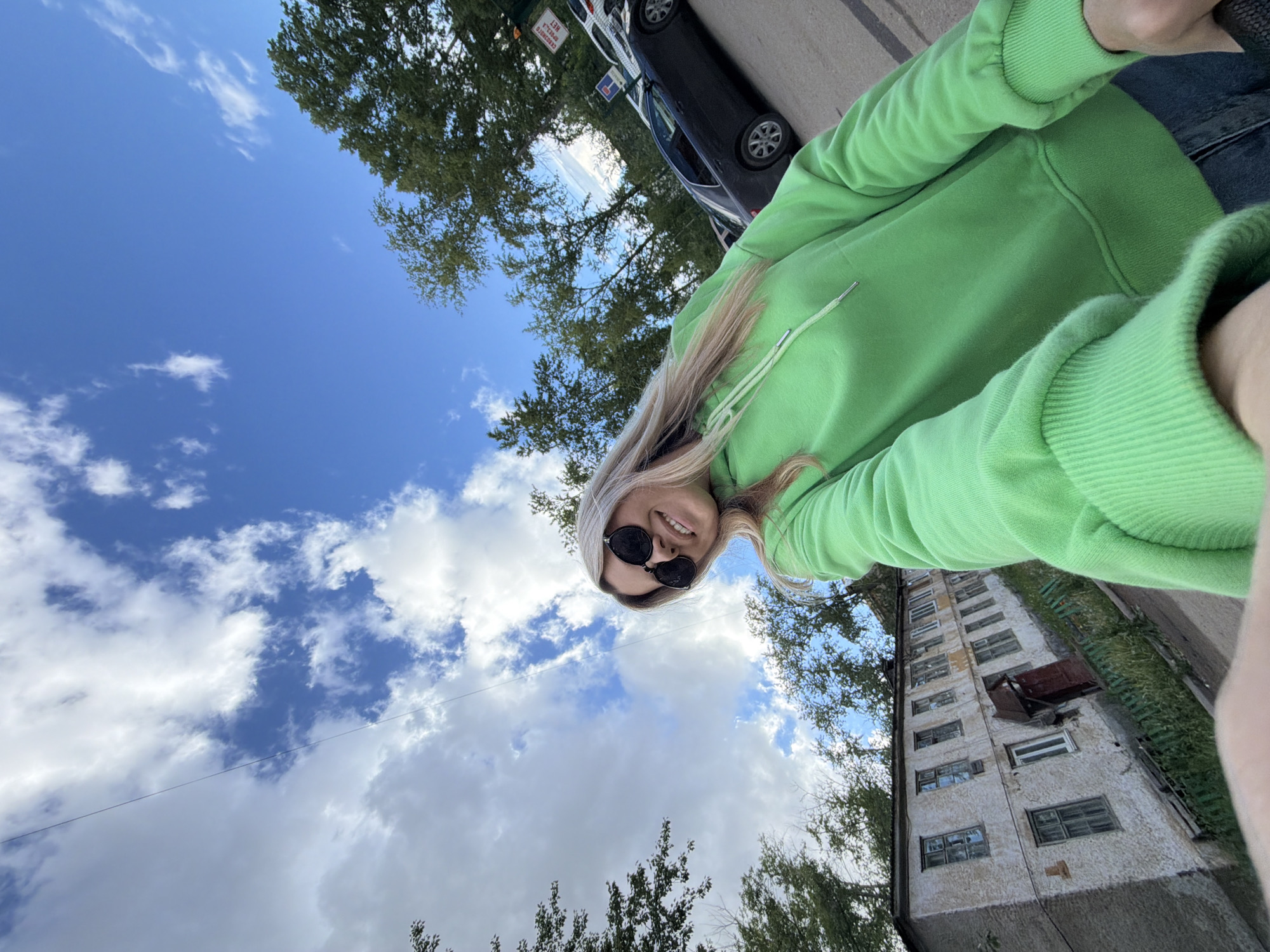 The image is a bright, high-angle selfie taken by a woman on a sunny day in Chaikovsky, Russia. The woman, positioned in the foreground and looking up at the camera, has long blonde hair and is wearing dark, oval-shaped sunglasses. She is smiling broadly and dressed in a vibrant, light green hooded sweatshirt, with a glimpse of blue jeans visible.
The photo's rotated perspective creates a dynamic view, as if taken from a low angle looking upwards. The background features a clear blue sky with scattered white clouds and the leafy canopies of several green trees. On the left side, a street is visible with a black car and a white car parked. A white sign with red Cyrillic text is partially visible near the cars, displaying "САМОРЕЗЫ" (screws) and "НЕТ" (no). To the right, a multi-story, possibly residential building with a textured, light-colored facade and numerous windows stands tall, appearing somewhat aged. The overall scene suggests a pleasant outdoor moment in an urban or suburban setting.