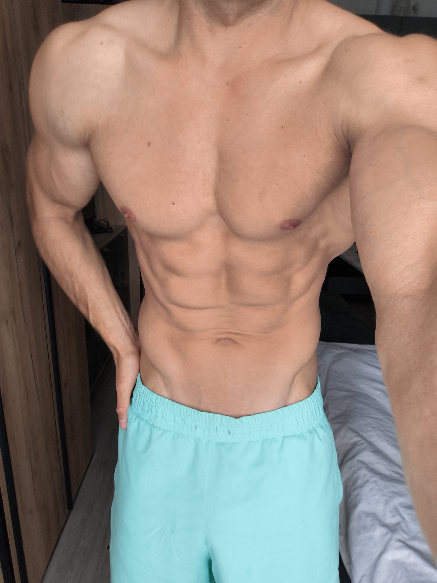 A shirtless male individual with a muscular build is seen in a room. The background features a wooden cabinet to the left and a bed with white sheets on the right. The man is wearing light blue shorts. The location cannot be determined to be Moscow, Russia based on the provided image.