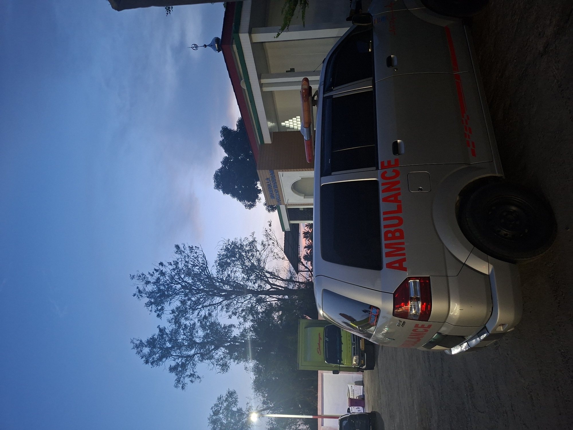 A silver ambulance is parked in an outdoor lot in front of a building that appears to be a mosque. The mosque has white walls, a red-trimmed roof, and a small dome. The ambulance has red lettering on its side that spells out "AMBULANCE". A green truck is parked behind the ambulance. Trees and a street light are visible in the background. The sky is a deep blue, suggesting it is either dawn or dusk. The location is identified as Sinyeu, Indonesia.