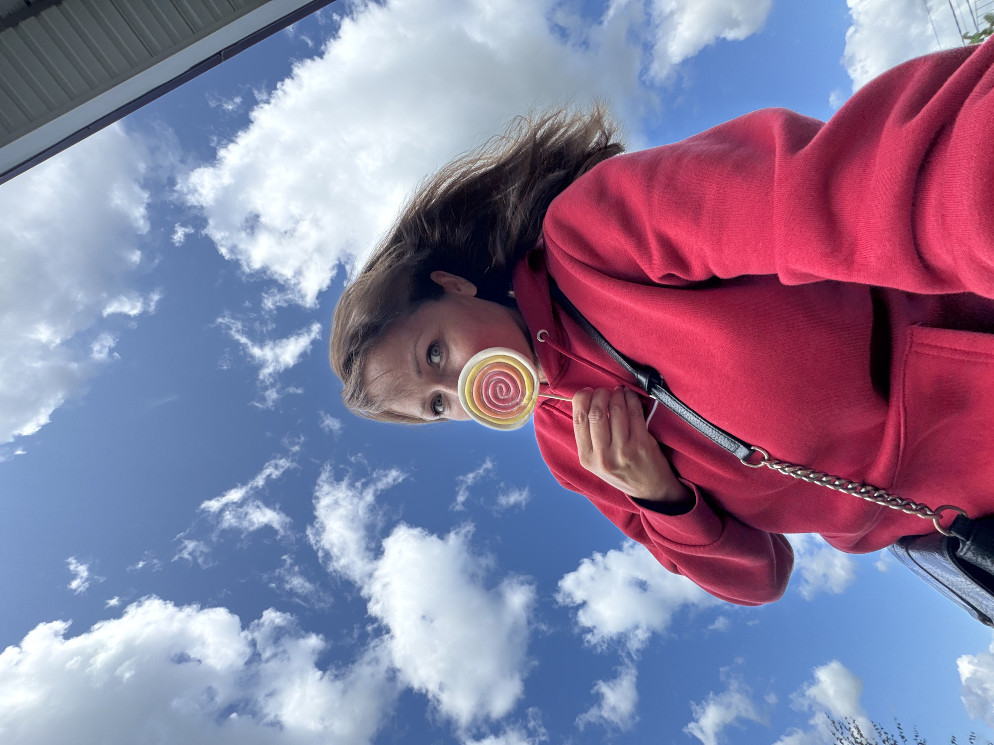 In Kempelevo, Russia, a woman is photographed from a low angle, holding a swirled lollipop in front of her mouth. She is wearing a red hoodie and has her hair blowing in the wind. The background is a bright blue sky filled with fluffy white clouds, with the edge of a building visible at the top right.