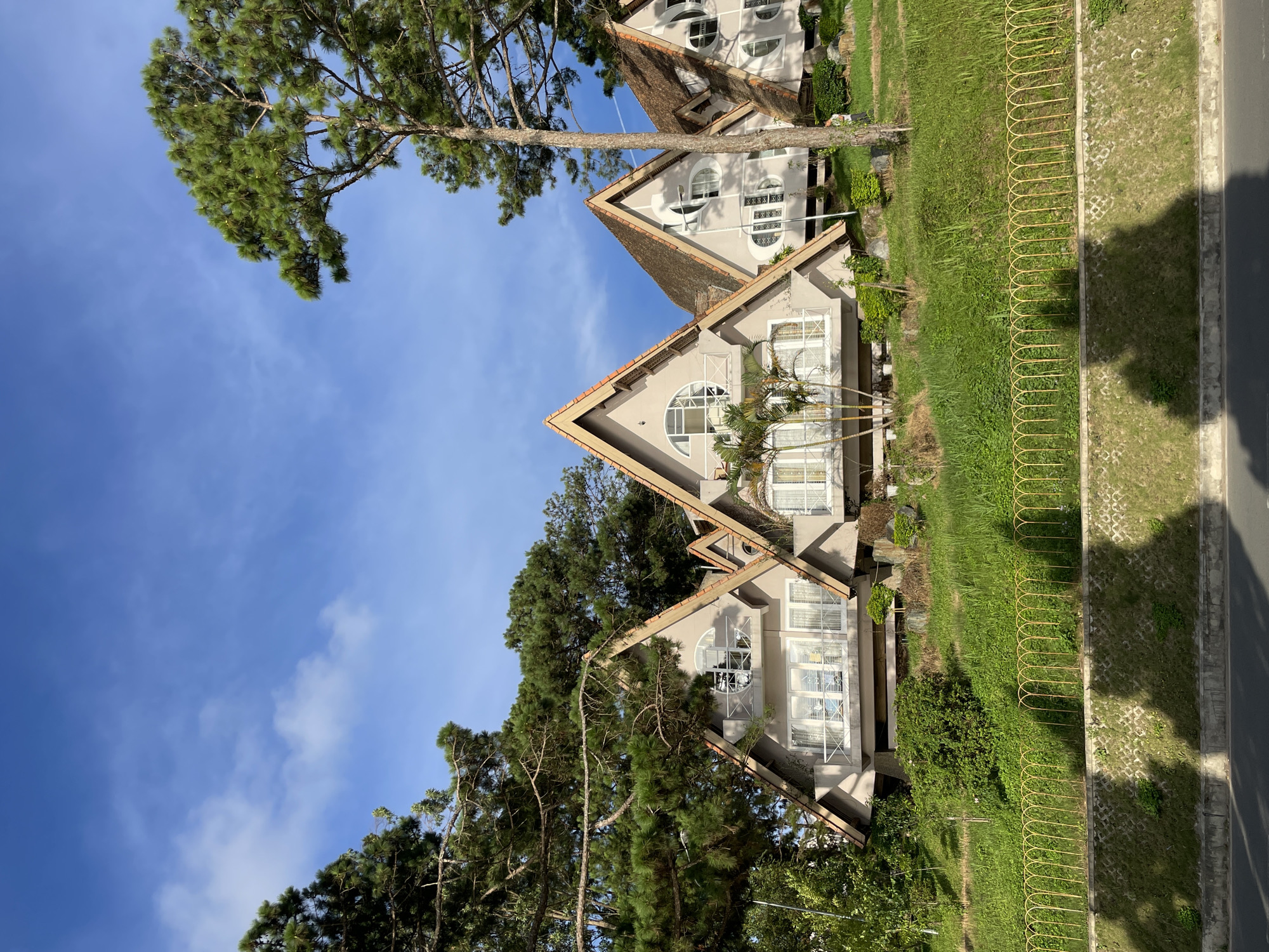 The image depicts a row of distinctive triangular-shaped houses situated on a hillside in Da Lat, Vietnam. The architecture of the houses is characterized by steep, gabled roofs and multiple levels with large windows and balconies. The houses are painted a neutral beige color and are nestled among lush green trees and vegetation, including tall pine trees. A yellow, arched fence runs along the foreground, separating the hillside from a paved road. The sky above is a clear blue with scattered white clouds, suggesting a pleasant day. The scene is tranquil and serene, conveying a sense of architectural charm and natural beauty.