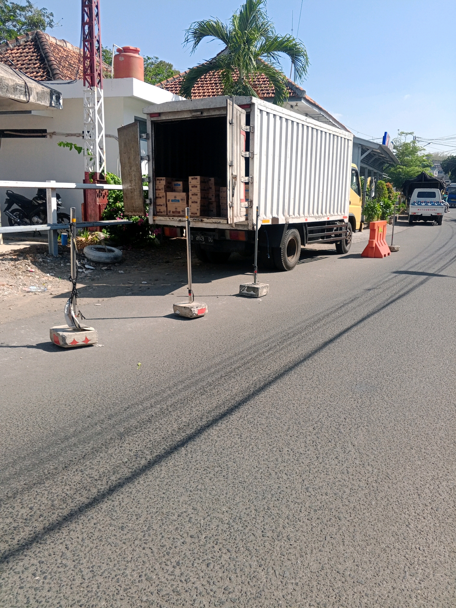 A bright, sunny day in Wonogiri, Indonesia, illuminates a scene on a paved road where a large white box truck is parked with its rear doors open, revealing stacks of cardboard boxes inside. The truck, a light yellow cab with a white cargo area, is positioned on the right side of the road. In front of the truck, an orange traffic barrier stands on the asphalt.
To the left of the truck, a motorcycle is parked near a white fence. Further left, a building with a red-tiled roof is visible, alongside a white metal tower structure supporting an orange water tank. Two portable traffic stanchions, each with a concrete base and a metal pole, are placed on the road in the foreground. The road itself has tire marks and shadows cast by the truck and the stanchions. In the distance, other vehicles are on the road, including a white pickup truck with "MITSUBISHI" written on its tailgate and several passengers. The environment suggests a typical urban or suburban street in Indonesia.