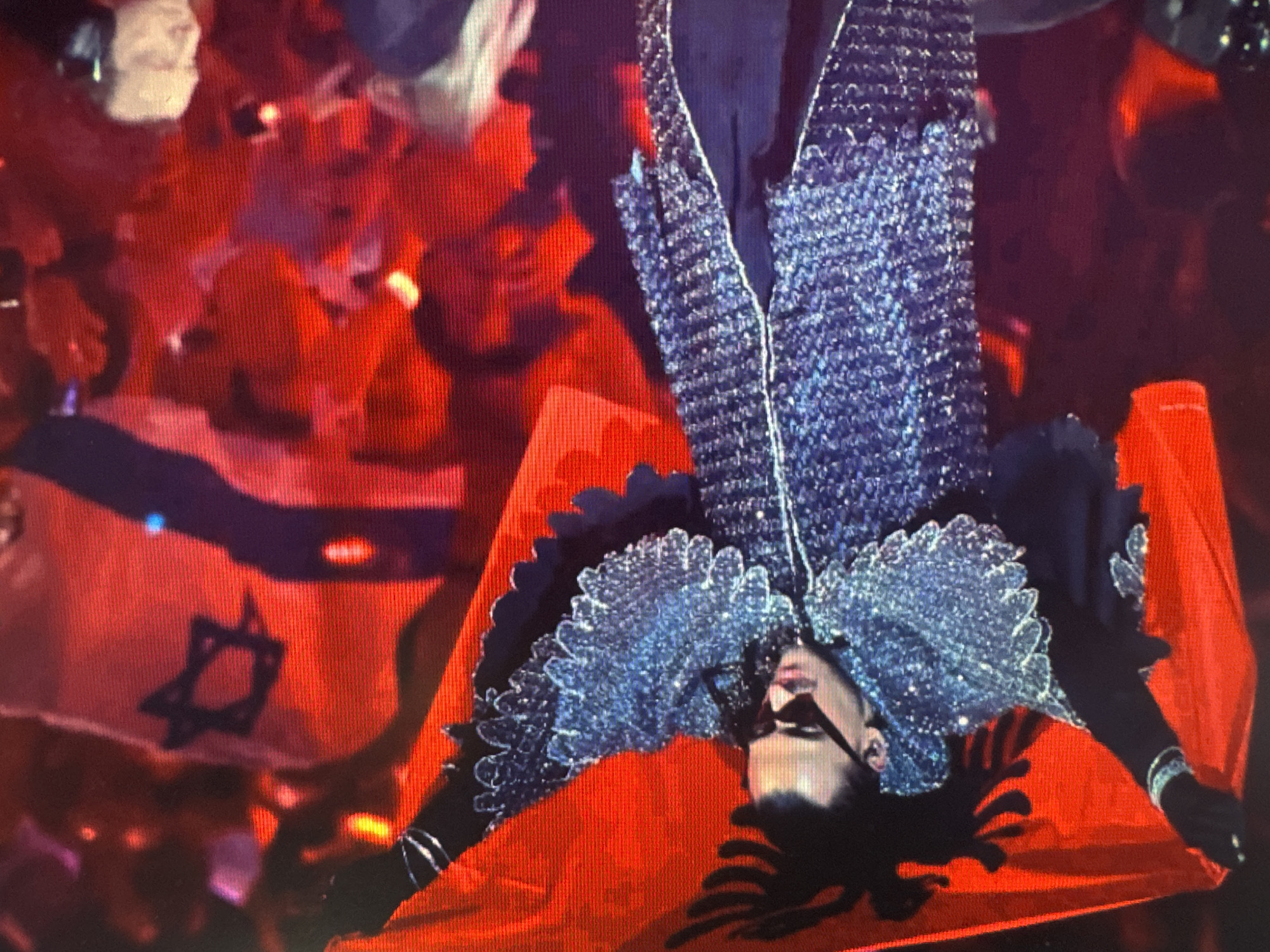 The image depicts a person in a highly decorative, sparkly outfit, holding two flags. The person is wearing dark sunglasses and has black hair. The outfit appears to be a jacket with wide, shimmering lapels and possibly a cape or train that extends downwards. The person is positioned upside down in the frame, with their head at the bottom.
One of the flags is predominantly red with a dark six-pointed star (Star of David) in the center and a horizontal blue stripe above it. This is the flag of Israel. The other flag is solid red, with a black double-headed eagle insignia. This is the flag of Albania.
The background is a blur of red and some lighter shapes, suggesting a crowd or audience. The overall impression is that of a performance or event, possibly a concert or a public gathering, where flags are being displayed. The lighting is dramatic, with bright highlights and deep shadows, creating a theatrical atmosphere. The context provided is Ellwangen, Germany, which could indicate the location of this event. The emotion conveyed is difficult to determine due to the sunglasses and the upside-down orientation, but the elaborate costume and the display of national flags suggest a celebratory or significant moment. There is no visible text in the image.