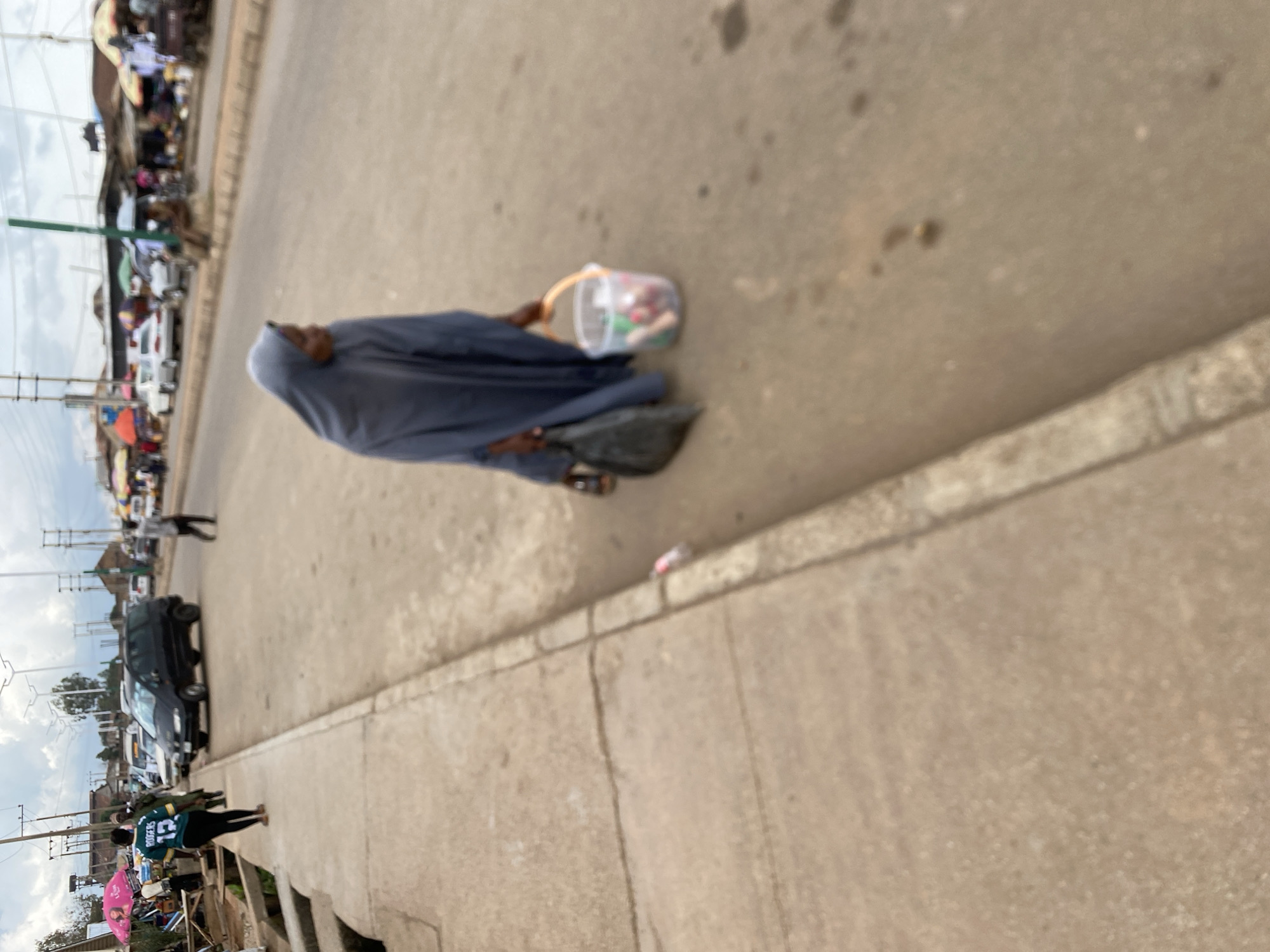 A street scene in Bukuru, Nigeria, features a person in a blue headscarf and garment walking on the right side of a paved road, carrying a black plastic bag in their left hand and a clear plastic bucket containing colorful items in their right hand. To the left, a paved sidewalk runs alongside the road, bordered by a low concrete structure. Several vehicles, including a black van, are visible on the road and in the background, along with market stalls and utility poles. The sky is overcast.