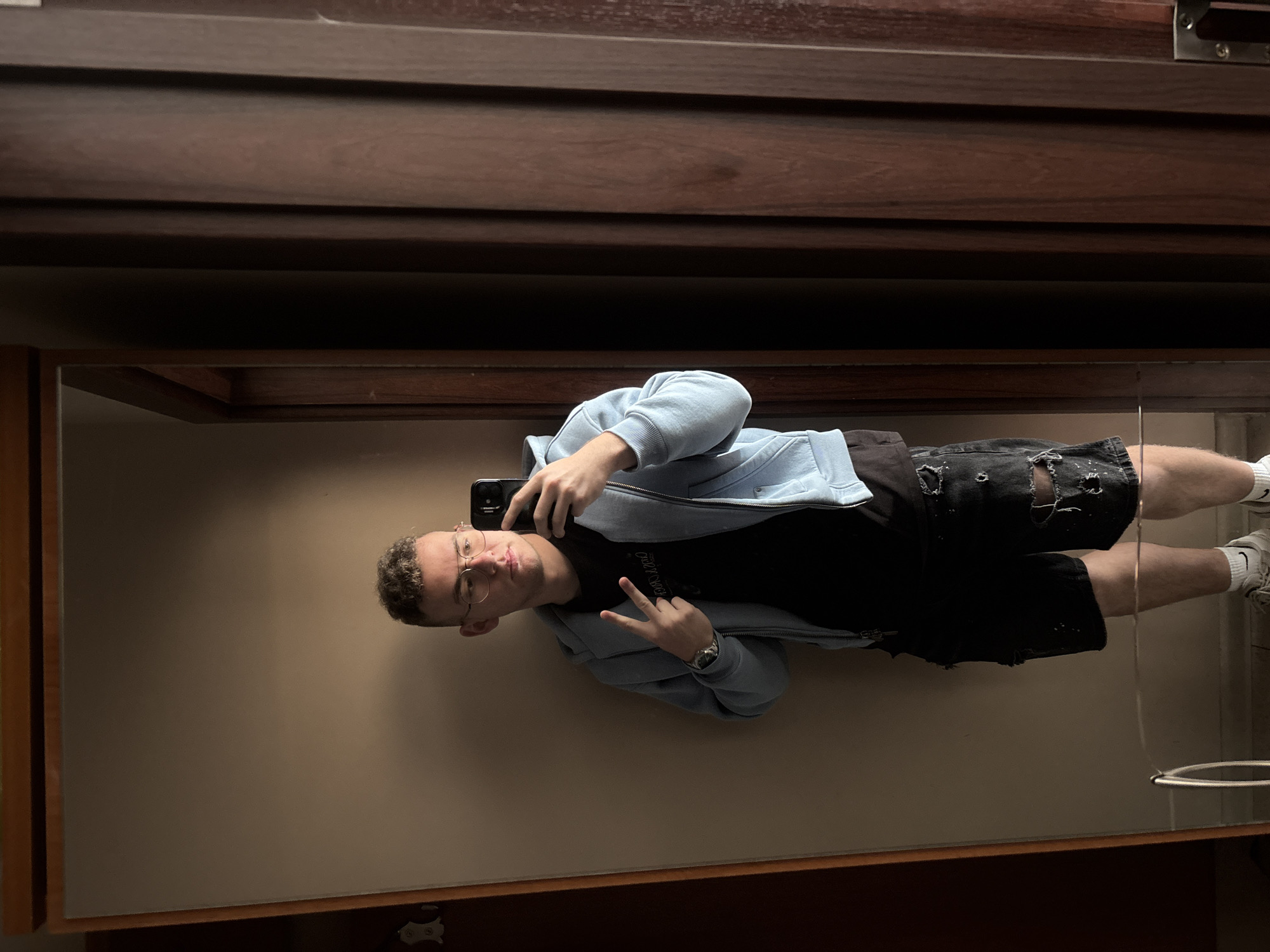 A young man stands in front of a large rectangular mirror with a dark wooden frame, taking a selfie. He is wearing round glasses, a black t-shirt, a light blue zip-up hoodie, and ripped black denim shorts. He is holding a black smartphone in his right hand, positioned to capture his reflection, and making a peace sign with his left hand. His sneakers are visible in the lower right corner. The mirror reflects a plain, light brown wall. Above the mirror is a dark brown wooden structure, possibly part of a dresser or wardrobe. The location is unknown and the city cannot be determined from the image.