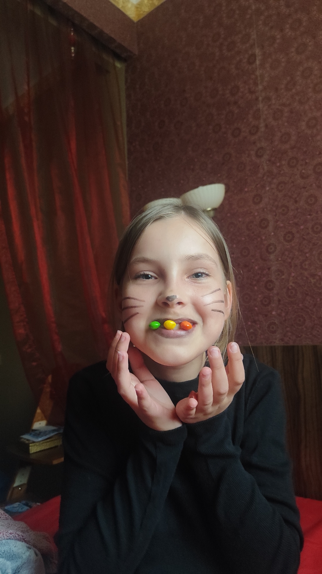 A young girl with blonde hair, cat-like face paint (whiskers and a nose), and three colored candies (green, yellow, and orange) held in her mouth smiles at the camera. She wears a long-sleeved black shirt and holds her hands near her face in a playful manner. The background includes a patterned brown wallpaper, part of a light fixture above her head, and a section of red curtains. To the left, there is a table or shelf with books and other items. The scene appears to be indoors, possibly a bedroom or living room. This picture was taken in Chaikovsky, Russia.