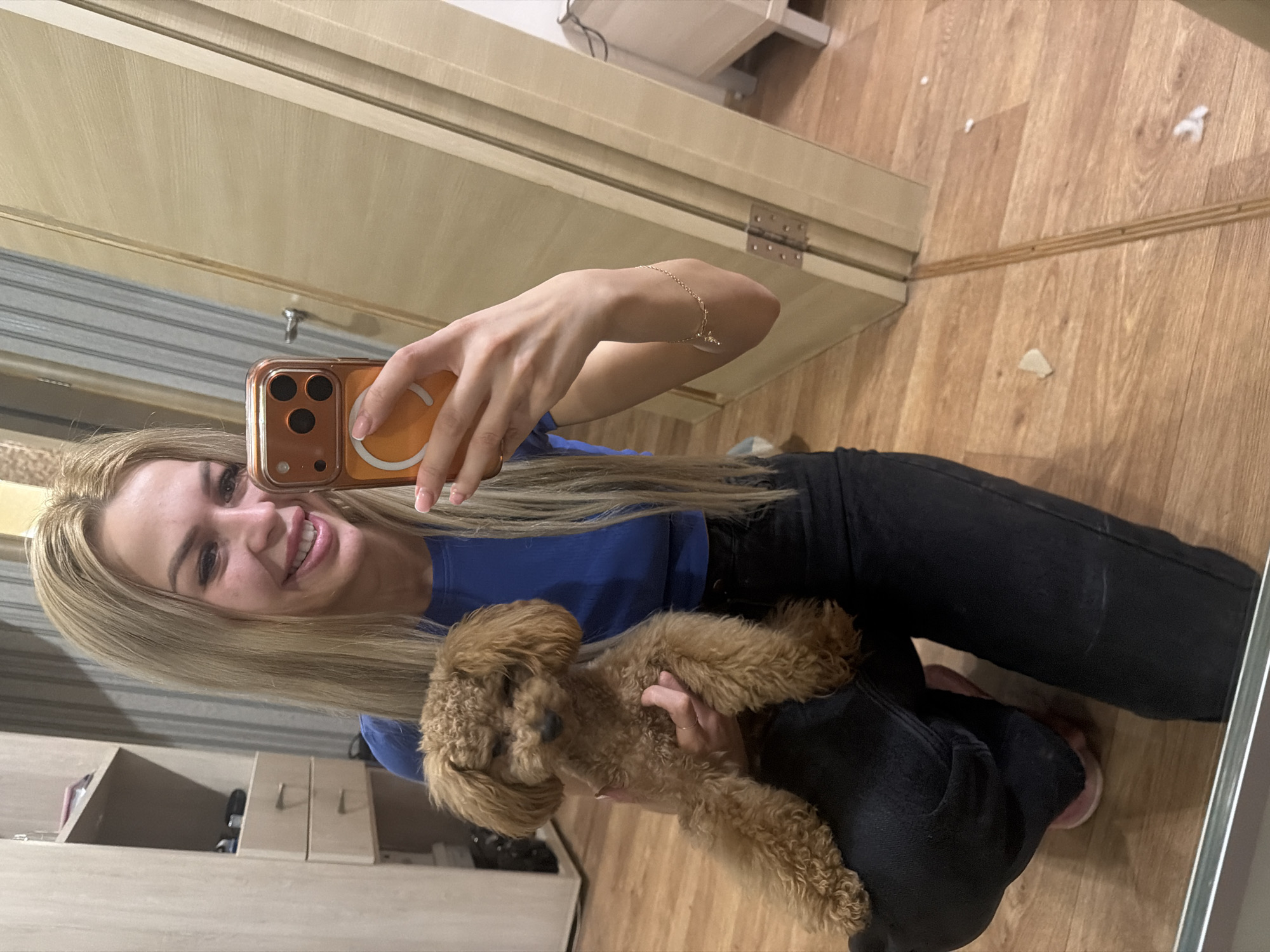 A young woman with long blonde hair and a blue shirt is taking a selfie in a room. She is holding a small, brown poodle in her arms. The poodle is looking down, and the woman is smiling at the camera. The room appears to be a bedroom or living room, with wooden floors and a wardrobe in the background. The woman is wearing black jeans and pink slippers. The selfie is being taken with a smartphone with an orange case. The background includes a partially open door and a mirror. The scene is indoors and appears to be a residential setting. There are no clear indicators of the specific location within Kyiv, Ukraine.