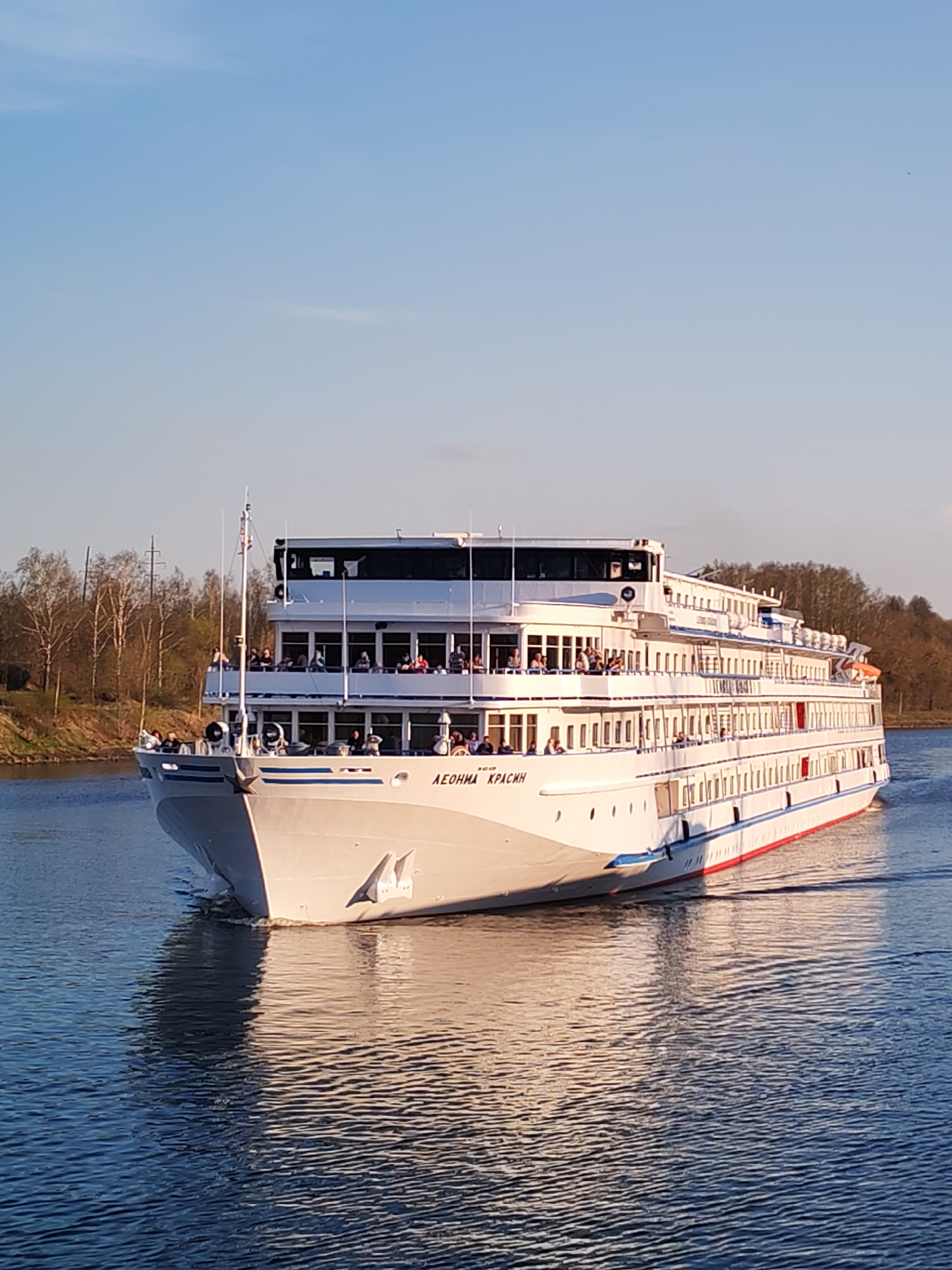 The image shows a large white passenger ship, named "Леонид Красин" in Cyrillic, traveling on a body of water. The ship has multiple decks and numerous windows, with passengers visible on the decks. The water reflects sunlight, creating a shimmering effect. The background consists of trees and vegetation along the shoreline. Based on the ship's name, Базарово is unlikely the location. It is more likely to be located on a major waterway in Russia, like the Volga River or a canal system.