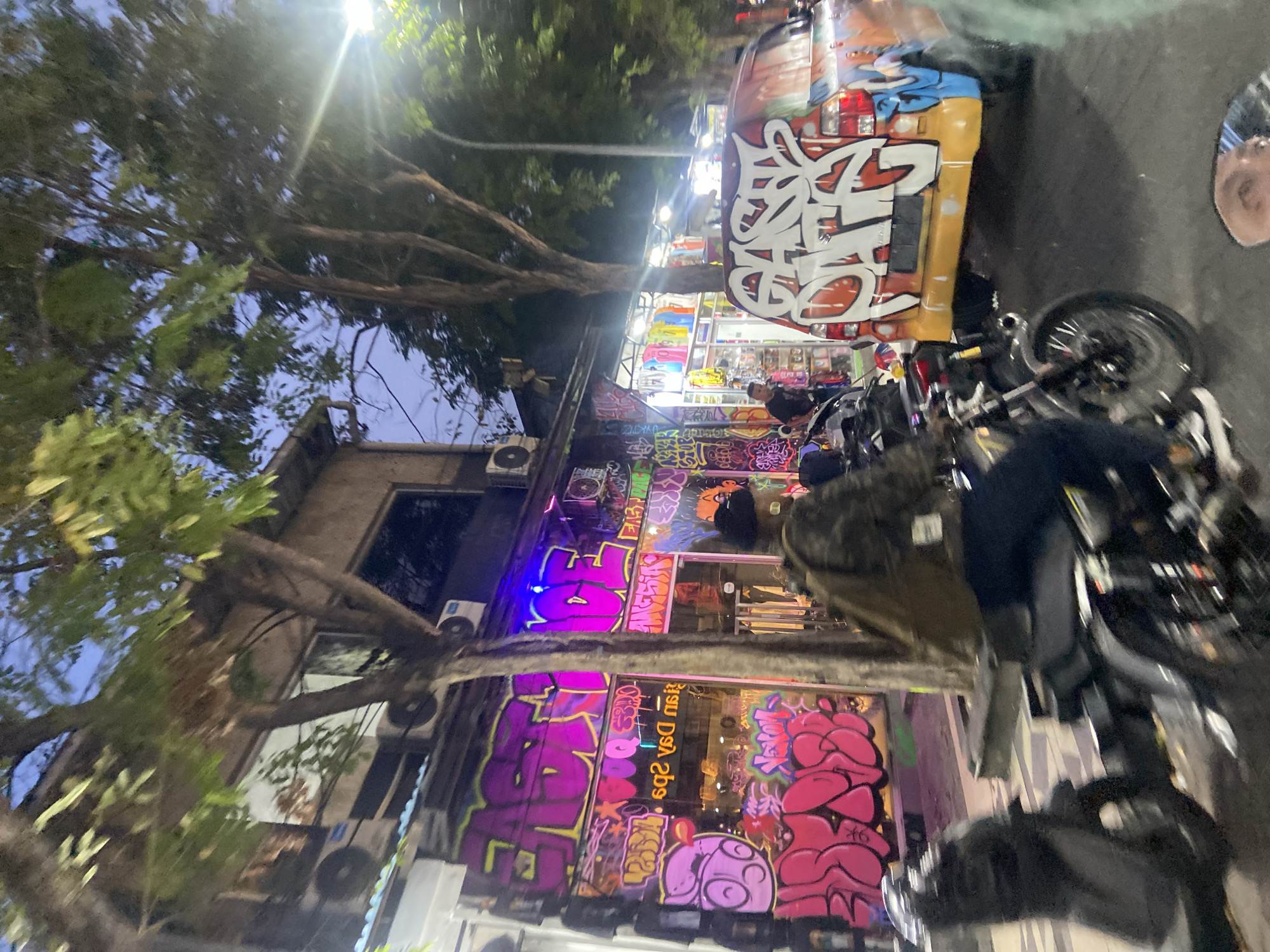 The image presents a vibrant and bustling night street scene, rotated 90 degrees clockwise, likely depicting a commercial area in Kuta, Indonesia. Bright artificial lights from shops and streetlamps illuminate the scene, creating prominent lens flares against a dark evening sky.
On the left, a multi-story building features ground-level shopfronts extensively covered in colorful and dynamic graffiti. One section prominently displays bold purple letters that appear to spell "LOVE," while other areas burst with stylized pink bubble letters, intricate tags, and various urban art designs. A shop window reflects inverted text, possibly a sign from inside or across the street, appearing as "qsə pSᗡ isŋ." Air conditioning units are visible on the building's facade.
Parked on the right side of the street are several motorcycles, a common sight in the region. Behind them, a customized vehicle, possibly a pick-up truck or van, stands out with its entire body covered in large, elaborate white and colored graffiti. A person in a dark jacket is partially obscured by one of the motorcycles in the mid-ground. The partial reflection of a person's face is visible in a vehicle mirror on the far right.
Large trees with dense foliage frame the top and left sides of the scene, their branches silhouetted against the night sky. The extensive street art, numerous motorcycles, and brightly lit shops collectively suggest a lively urban environment, indicative of a popular tourist or commercial district active during the evening. The atmosphere is energetic and visually rich.