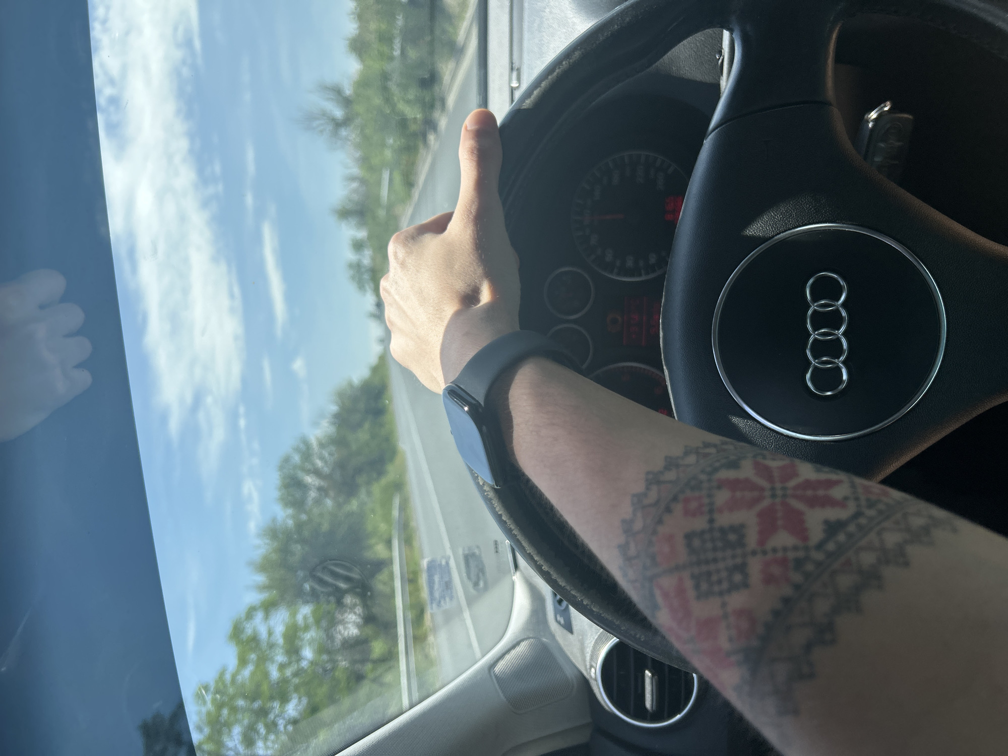 The image captures a first-person perspective from inside a car, showing a person's right arm and hand giving a thumbs-up gesture while driving. The person is wearing a smartwatch on their wrist, and their forearm features a prominent tattoo with a geometric, embroidery-like pattern in red and dark gray/black. The steering wheel, bearing the distinctive Audi logo of four interconnected rings, is central to the foreground. Behind the steering wheel, parts of the car's dashboard and instrument cluster are visible, showing faint red indicators.
Through the car's windshield, a sunny outdoor scene is visible. The road stretches ahead, flanked by green trees. The sky is bright blue with scattered white clouds, suggesting clear weather during the daytime, likely midday or afternoon given the strong sunlight and shadows. Reflections of the sky and clouds are also visible on the car's rearview mirror. The thumbs-up gesture conveys a positive sentiment, possibly satisfaction or enjoyment of the journey. The setting is Mihiltsi, Bulgaria, indicating the location of this drive. No other decipherable text is visible besides the Audi logo and the patterned tattoo.