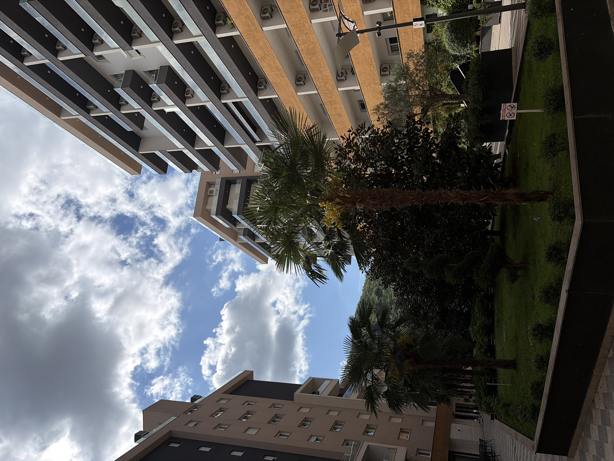 An outdoor scene in Budva, Montenegro, captures a modern urban landscape from a low-angle perspective, looking up towards the sky and surrounding buildings. The dominant subjects are contemporary apartment or hotel buildings, lush greenery, and a partly cloudy blue sky.
Two multi-story buildings frame the view. The building on the left features a striking design with dark, horizontal bands alternating with lighter sections, likely balconies or architectural elements, some appearing to be glass-fronted. Numerous external air conditioning units are visible along the building's facade. Another building, slightly lighter in color, is seen on the right side of the image, also with balconies and multiple windows.
Between and in front of the buildings, a vibrant green space is visible, characterized by several palm trees with distinctive, textured trunks and fronds. Other dense green trees and bushes contribute to the verdant environment, suggesting a well-maintained garden or courtyard. In the background, a large, green, tree-covered mountain rises, typical of coastal Montenegro.
The sky is bright, indicating daytime, with scattered white cumulus clouds against a clear blue expanse. The overall lighting suggests a sunny day.
No people are visible, and no specific activity is taking place; the scene is static, showcasing the architecture and natural elements.
Several signs are present in the green area. One white sign with red borders clearly shows two crossed-out symbols: a dog, indicating "no dogs," and a cigarette, indicating "no smoking." Below these symbols, Italian text reads "VIETATO FUMARE" (No Smoking) and "NON CALPESTARE L'ERBA" (Do not step on the grass). Another sign with similar symbols is partially visible nearby. The scene conveys a sense of modern development integrated with nature in a clean, regulated environment.