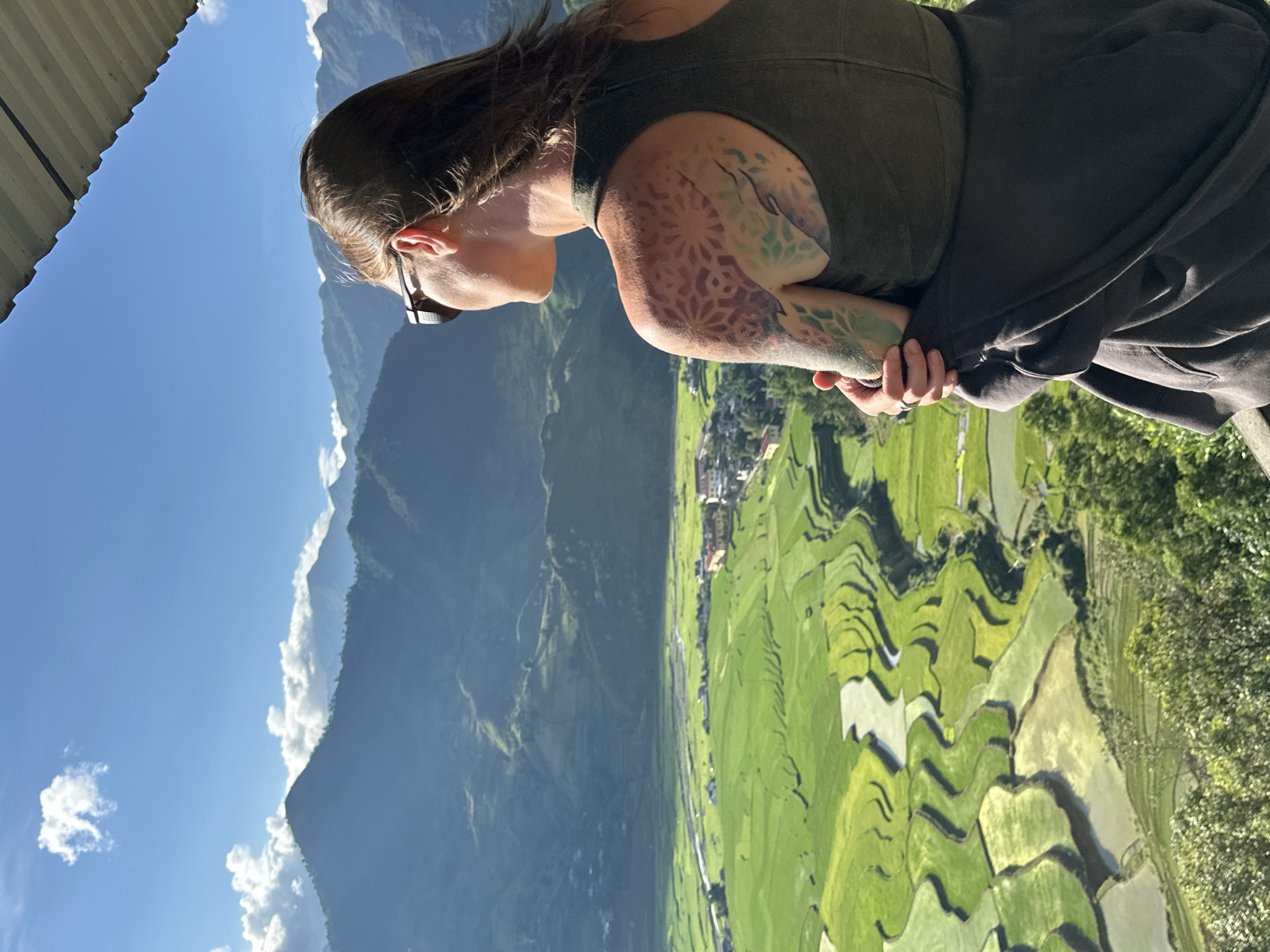 A person is observed from the back, facing away from the camera, looking out over a verdant valley in Tú Lệ, Vietnam. The individual, a female, wears dark sunglasses, a dark olive green tank top, and has a dark gray jacket tied around her waist. A detailed, colorful tattoo featuring geometric and organic patterns is visible on her left shoulder and upper arm. Her brown hair is parted on the left and falls over her shoulders.
The expansive landscape before her consists of rolling mountains under a bright blue sky with scattered white clouds. In the foreground and midground of the valley, extensive terraced rice fields display varying shades of bright green and pale, water-filled sections, indicating different stages of cultivation. A river winds through the valley, passing near a cluster of buildings that form a small village or town. The sunlight casts clear shadows, highlighting the contours of the mountains and the intricate patterns of the rice paddies. The top right corner of the image shows a portion of a corrugated metal roof or overhang.