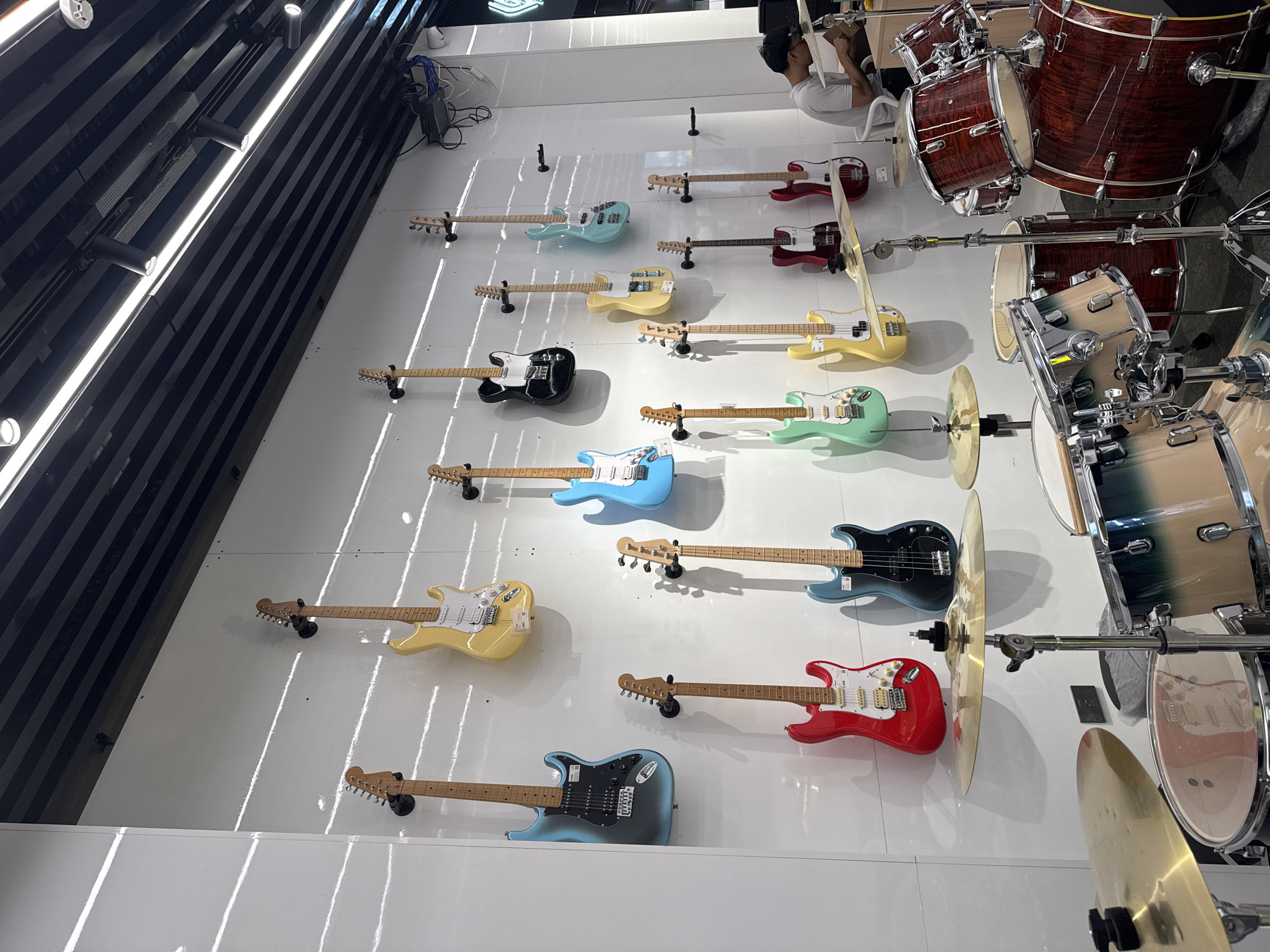 The image, captured from an elevated, downward-angled perspective, depicts an indoor scene likely within a music store or showroom in Davao City, Philippines. It showcases a vibrant display of musical instruments against a backdrop of clean, reflective white walls that strongly reflect bright overhead artificial lighting, creating striking vertical light streaks.
To the left, a dark, slatted wall section with modern track lighting fixtures adds a contrasting element to the bright display area. The main subjects are numerous electric guitars and a bass guitar, meticulously mounted on the white wall. These include multiple Fender Stratocaster-style guitars in colors such as light blue, yellow, red, and light green, along with a darker blue/gray model. Two Fender Telecaster-style guitars, one black and one light blue, are also visible, alongside a light blue bass guitar. The instruments are arranged in neat rows, presented as a retail or exhibition display.
On the right side of the image, two elaborate drum kits are prominently featured. The foreground drum kit has a rich, reddish-brown wood grain finish, complete with a kick drum, snare, tom-toms, and several cymbals (hi-hat, crash/ride). Behind it, another drum kit with a blue/green burst finish is partially visible. The associated drum hardware, including stands and mounts, is clearly seen.
In the upper right corner, a person wearing a white t-shirt is partially visible, appearing to be seated on a stool or chair, somewhat obscured by the drum kit. They seem engaged in an activity, possibly working on the instrument setup or inspecting the area. A tangle of electrical cables is also noticeable in this corner. The scene conveys a clean, well-lit environment focused on musical equipment, with no discernible text on any of the objects or surfaces.