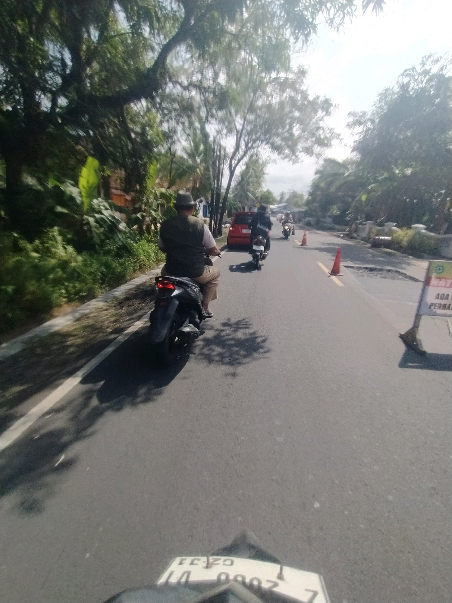 The image depicts a street scene in Desa Karangbenda, Indonesia, on a sunny day. The viewpoint is from the perspective of a rider on a motorcycle, evidenced by the front of a motorcycle and its license plate, "UZ-33.DUI.000Z," visible at the bottom of the frame.
In the foreground, a dark-colored motorcycle is in motion, with its rider wearing a light-colored shirt, a dark vest, light-colored pants, and a hat. The motorcycle is on the right side of the road.
Further down the road, other vehicles are visible. A red car is parked on the left side, and a motorcycle with a rider is approaching from the distance. Orange traffic cones are placed on the road, indicating a section of road work or a hazard, as a sign with Indonesian text, including "ADA PERBA..." (likely "ADA PERBAIKAN" meaning "There is repair"), is positioned on the right side of the road.
The environment is characterized by lush green vegetation and trees lining both sides of the road. Buildings are partially visible behind the foliage. The sky is bright, suggesting clear weather. The road itself appears to be asphalt, with white lane markings. The overall impression is that of a traffic scene in a semi-rural or suburban area.