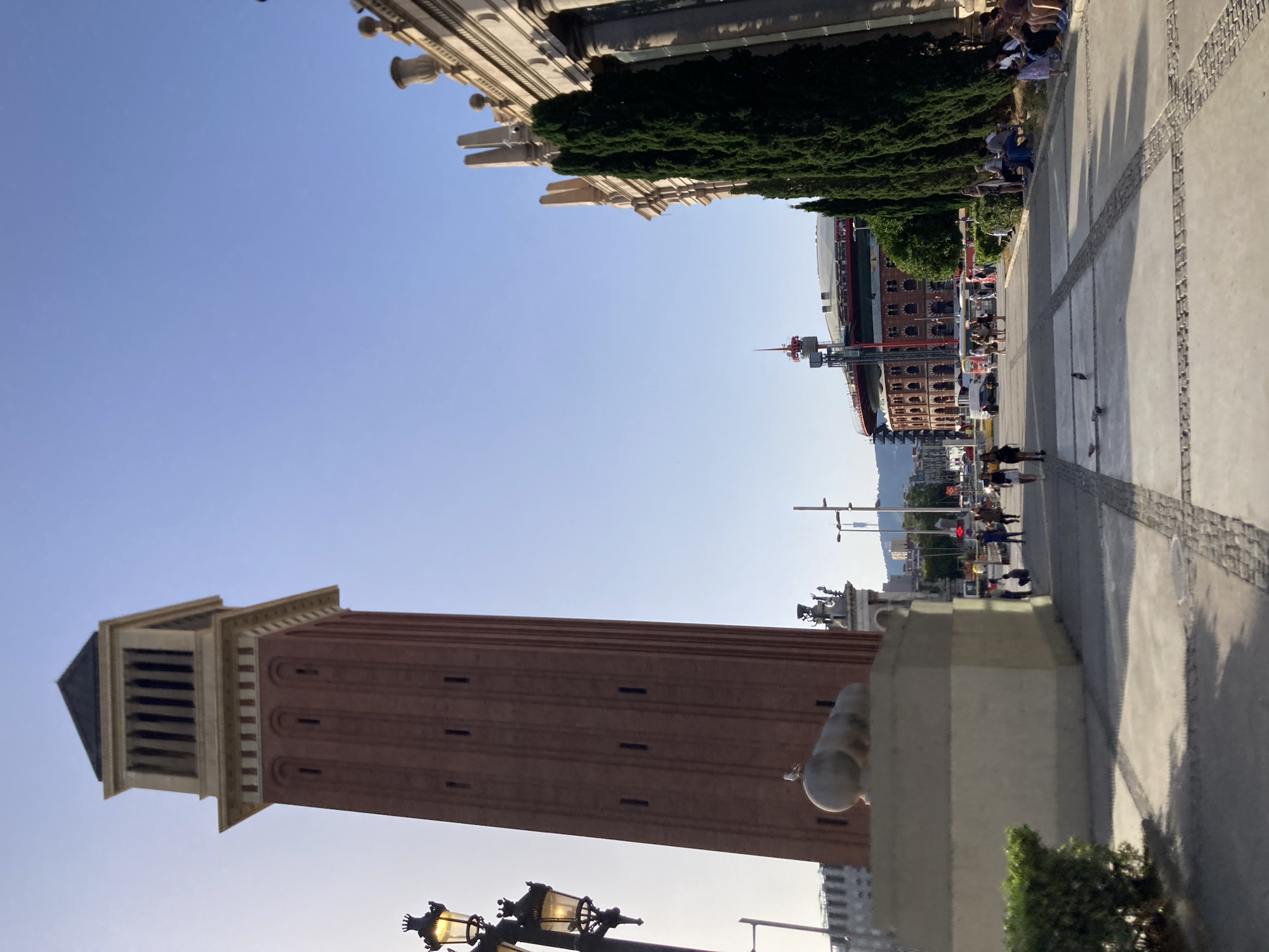 The image depicts the area in front of the Arenas de Barcelona, a former bullring converted into a shopping center, located on Gran Via de les Corts Catalanes in Barcelona, Spain.
In the foreground, a paved walkway with a patterned surface of square concrete slabs and cobbles leads towards the background. On the left side of the walkway, a tall, red brick tower with arched windows and a light-colored top section dominates the scene. A statue or monument, appearing to be a large, light-colored sphere supported by a rectangular base, is positioned at the foot of the tower. A bird is perched on top of the sphere. To the left of the tower, a decorative lamppost with multiple lights is visible.
To the right of the walkway, a large building with classical architecture, featuring columns and decorative urns, is partially visible. A row of tall, dark green cypress trees stands between this building and the walkway.
In the mid-ground, the circular structure of the former bullring, now the Arenas de Barcelona, is prominent. It is a red brick building with arches, topped by a modern red structure with a spired antenna. People are seen walking on the walkway and sitting on benches to the right. Several vehicles, including cars and buses, are present in the background, indicating an active urban environment. The sky is clear and blue, suggesting a daytime scene.
