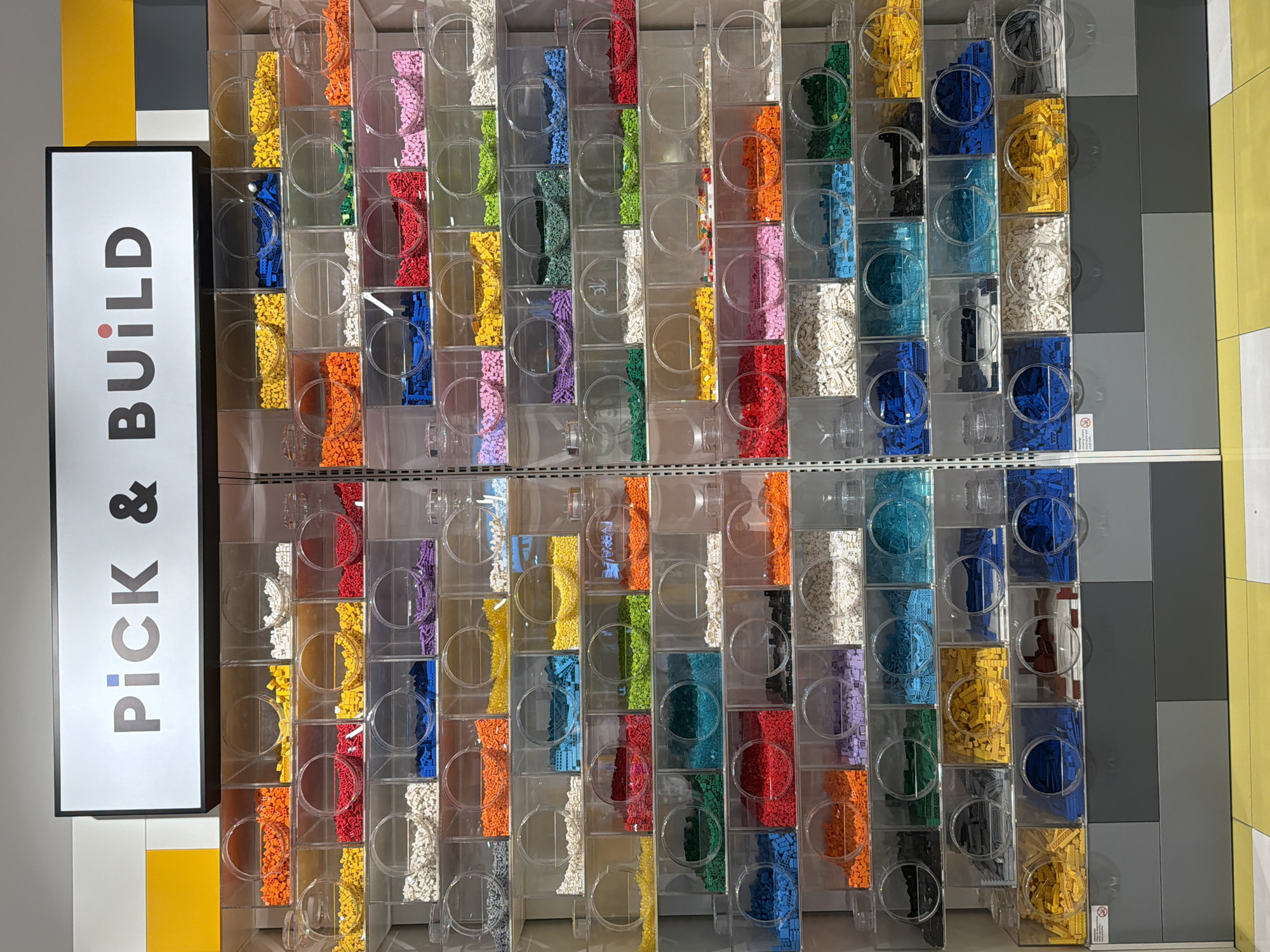 A "Pick & Build" Lego wall in a store in Puebla, Mexico, shows transparent bins arranged in a grid pattern. The bins are filled with Lego bricks of various colors including orange, white, red, purple, yellow, green, teal, blue, and black. A large white sign with black text that reads "PICK & BUILD" is visible to the left of the Lego wall. The surrounding wall has alternating panels of yellow and gray.