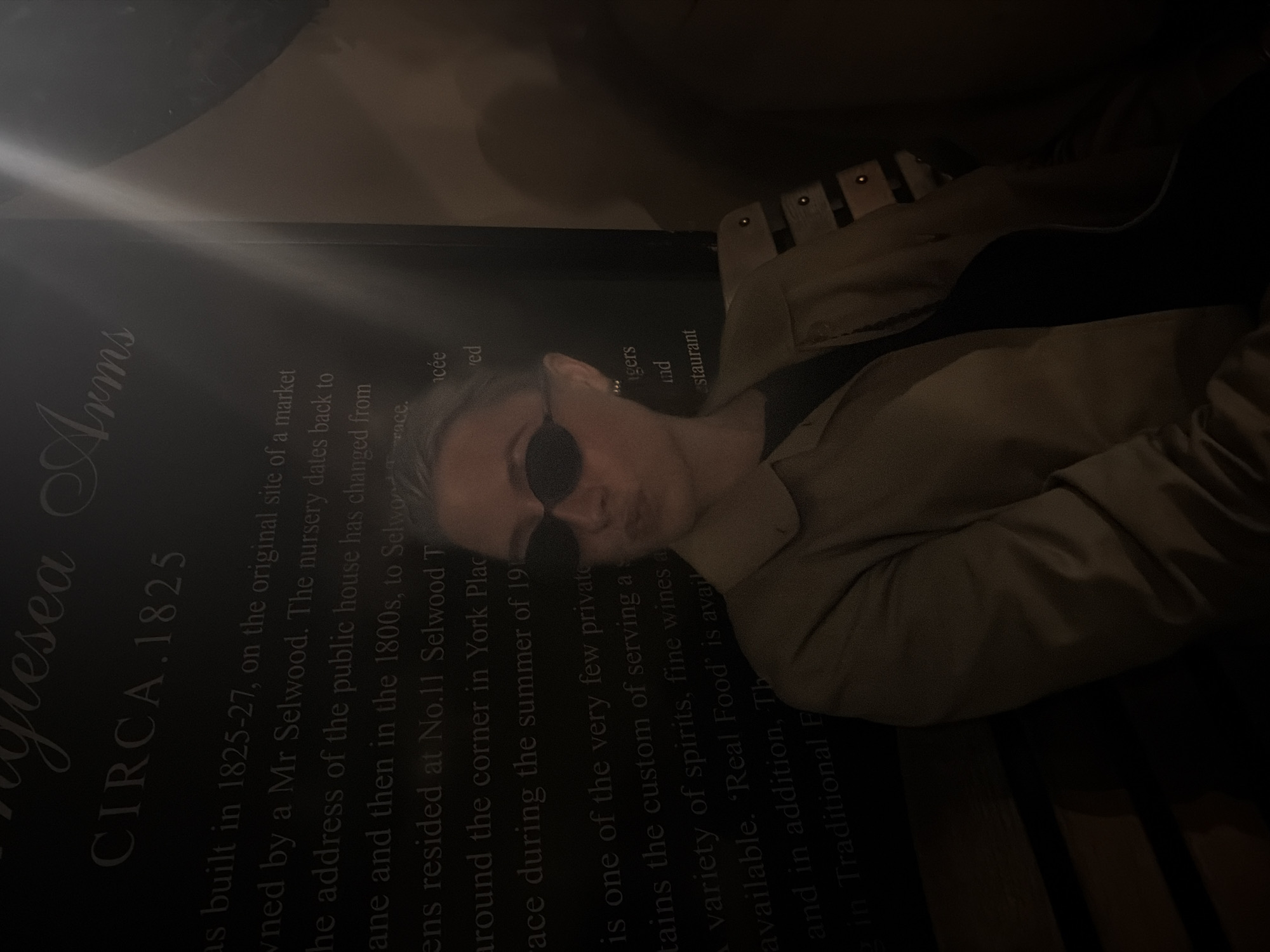A person wearing sunglasses and a beige jacket is leaning against a plaque with text that reads "Anglesea Arms CIRCA. 1825". The plaque is located in London, United Kingdom, based on the text mentioning "Selwood Terrace" and "York Place". The text describes the history of the Anglesea Arms, stating it was built in 1825-27 on the site of a market, once owned by Mr. Selwood, and the address changed from "lane" to "Selwood Terrace". The text also mentions the location near "No. 11 Selwood Terrace" and around the corner from "York Place," and that the establishment "maintains the custom of serving" a variety of drinks and 'Real Food'. The lighting in the image is low.