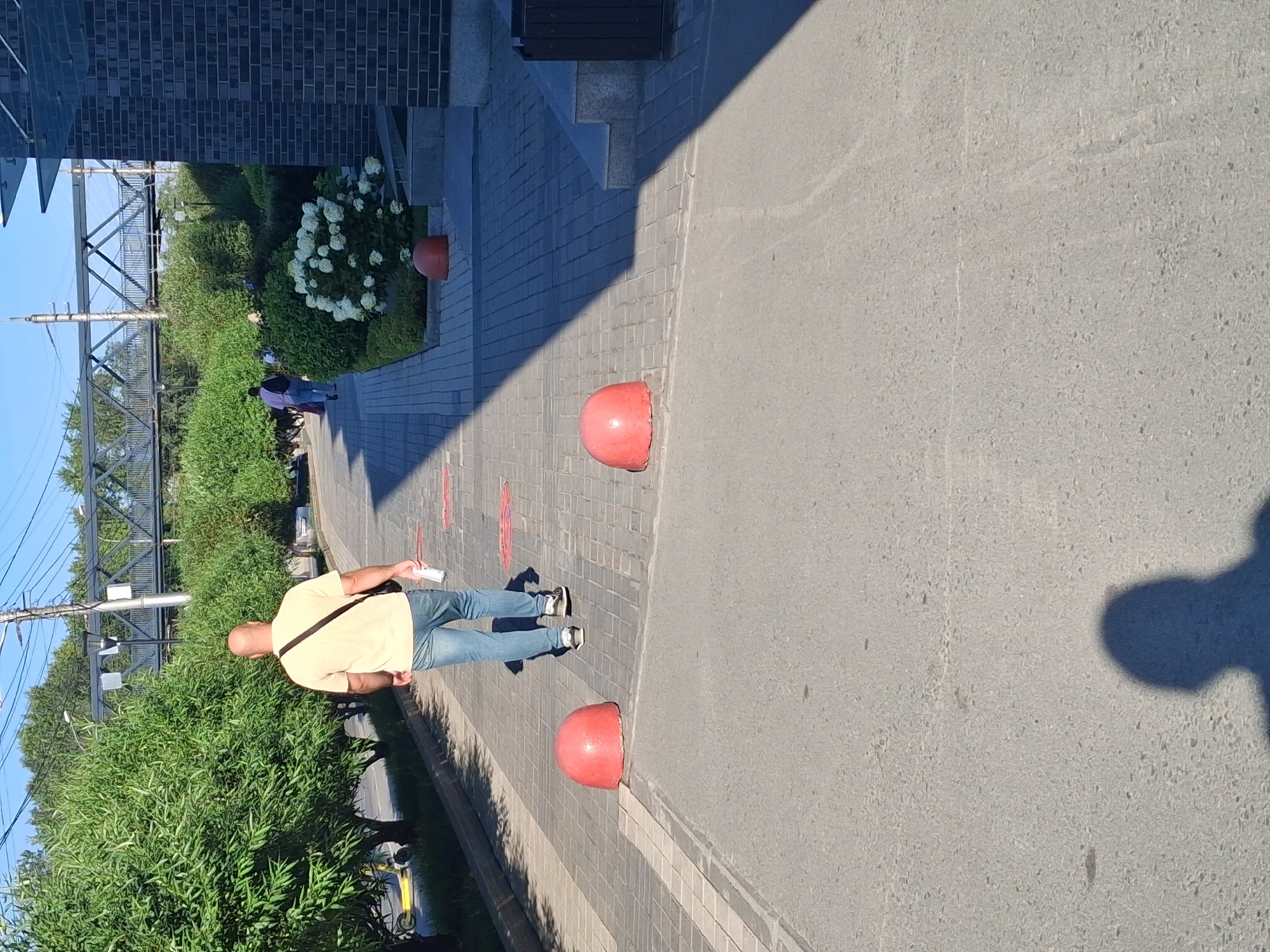 A bald man wearing a light-colored t-shirt and blue jeans walks away from the camera on a paved sidewalk. He carries a white cup in his right hand and a dark bag slung over his left shoulder. Two red, dome-shaped bollards are visible on the sidewalk ahead of him, partially in shadow. To the left of the sidewalk, a row of lush green trees lines the area, with a bright yellow electric scooter parked near the trees. In the background, a pedestrian bridge spans across a road or railway line, with overhead power lines visible. Further to the right, the corner of a dark brick building is visible, with steps leading up to it. Several people are walking on the sidewalk in the distance. The scene appears to be a public outdoor area in a city, possibly a park or a commercial district. The lighting suggests it is daytime, with strong shadows cast by the sun. The image is taken from a low angle.