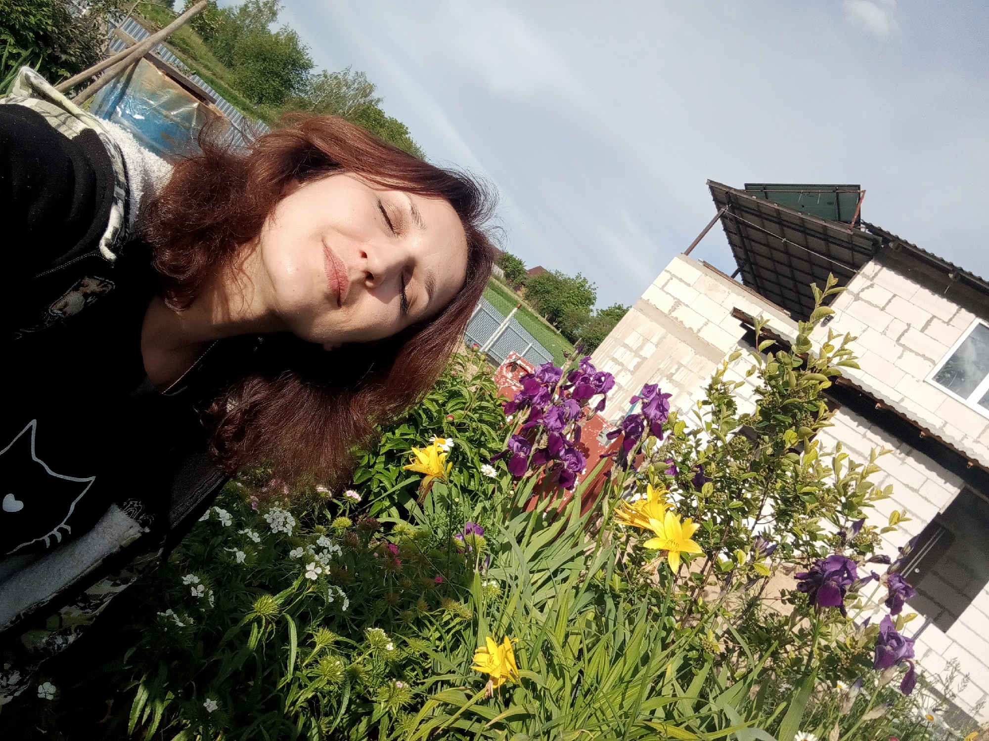 A woman with auburn hair is enjoying a sunny day in Babyn, Ukraine. Her eyes are closed as if basking in the warmth. She is wearing a black top with a white cat design. A variety of flowers in yellow, white, and purple are in the foreground, suggesting a garden. A partially constructed building made of white bricks is visible in the background, along with other houses and green trees under a bright sky. The scene conveys a sense of peace and tranquility.