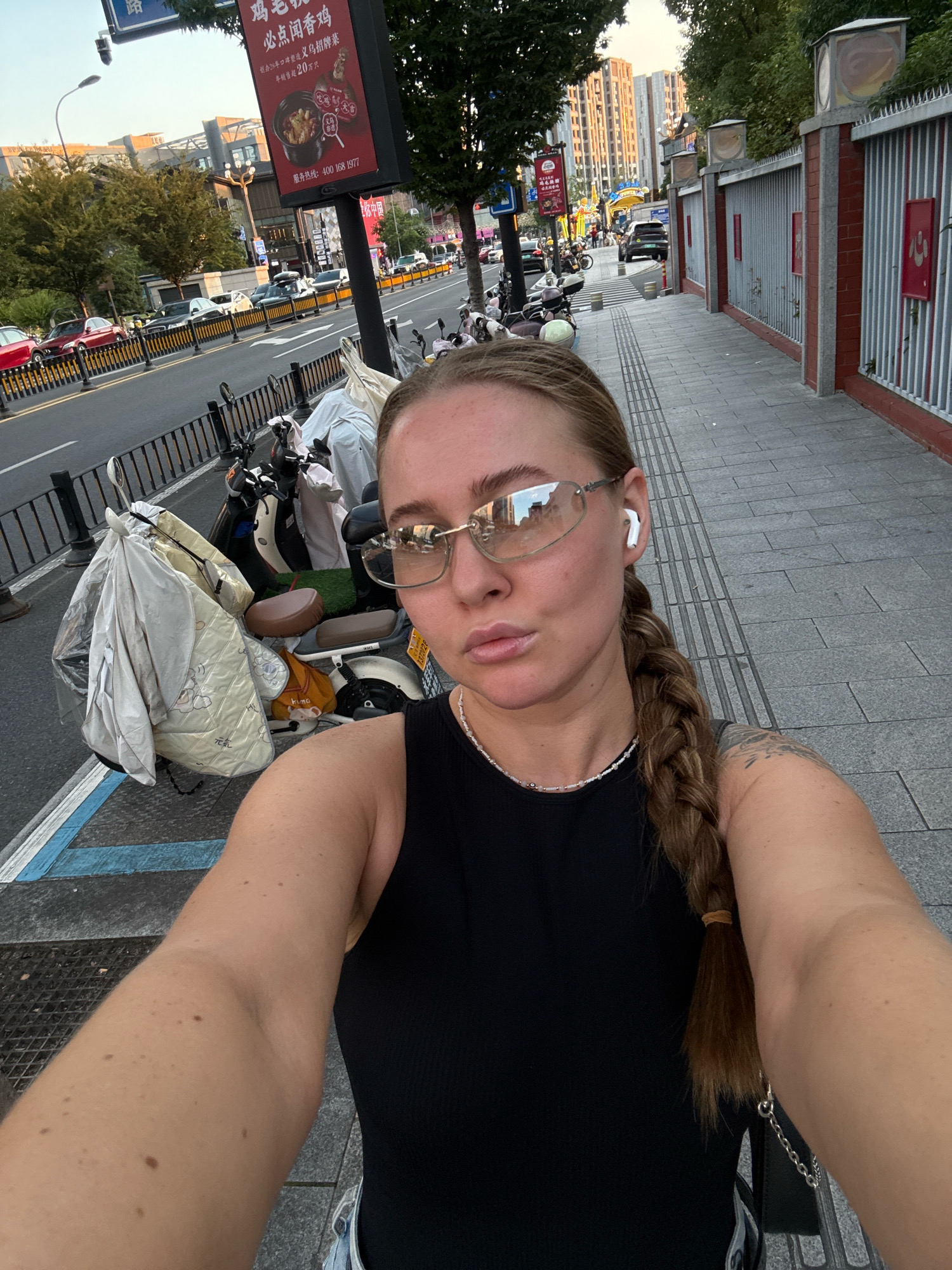 A woman is taking a selfie on a sidewalk in Yiwu, China. She has blonde hair in a braid and is wearing a black tank top, glasses, and white earbuds. In the background, there is a street with cars and motorcycles, as well as buildings and a red fence. The scene appears to be during the day, possibly late afternoon, given the lighting.