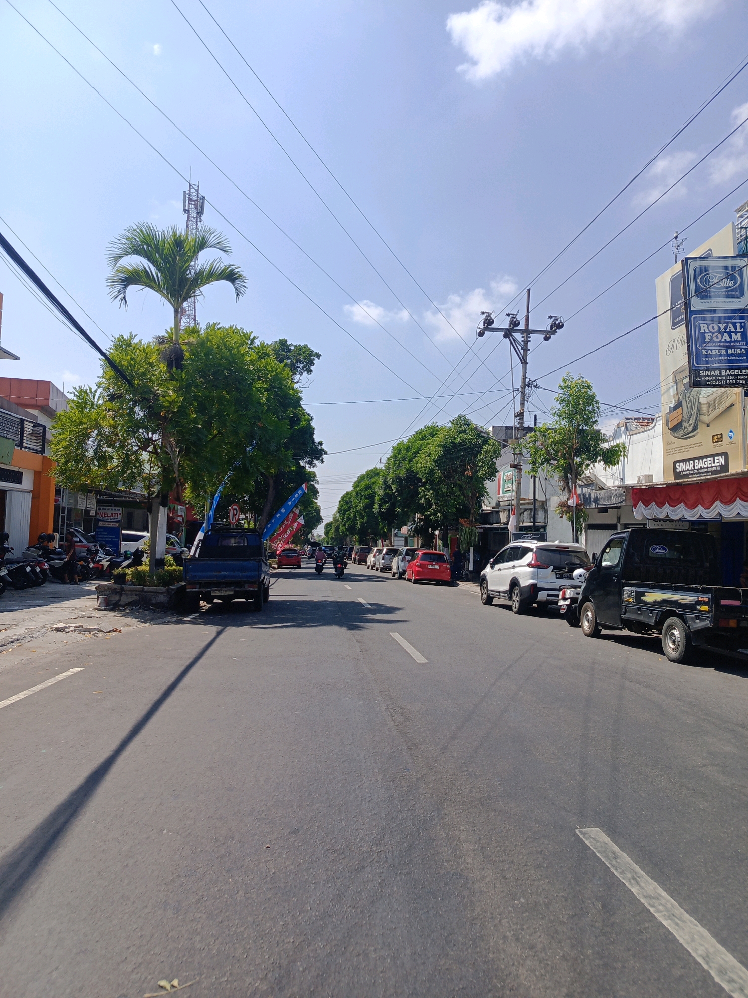 A street scene in Magetan, Indonesia, is captured on a bright, sunny day with a clear blue sky and scattered white clouds. The perspective is from the street, looking forward along a road with traffic.
On the left side of the street, there are several buildings with orange and white facades, and a sidewalk lined with parked motorcycles. Some trees provide shade along the sidewalk. A blue pickup truck is parked on the side of the road, with some blue flags attached to it.
The road itself is paved and appears to be in good condition, with white lane markings. Several cars and motorcycles are visible driving on the road. In the foreground, a shadow of a utility pole stretches across the asphalt.
On the right side of the street, more buildings are visible, including one with a large sign that reads "ROYAL FOAM" and "SINAR BAGELEN". A black pickup truck is parked in front of this building. Trees line the right side of the road as well, creating a canopy effect further down the street.
A prominent feature in the background is a tall cell phone tower, with a palm tree and other greenery at its base. Overhead, multiple power lines crisscross the sky, supported by utility poles. The overall impression is of a typical urban street scene in a Indonesian town, with a mix of commercial establishments, residential buildings, and everyday traffic.