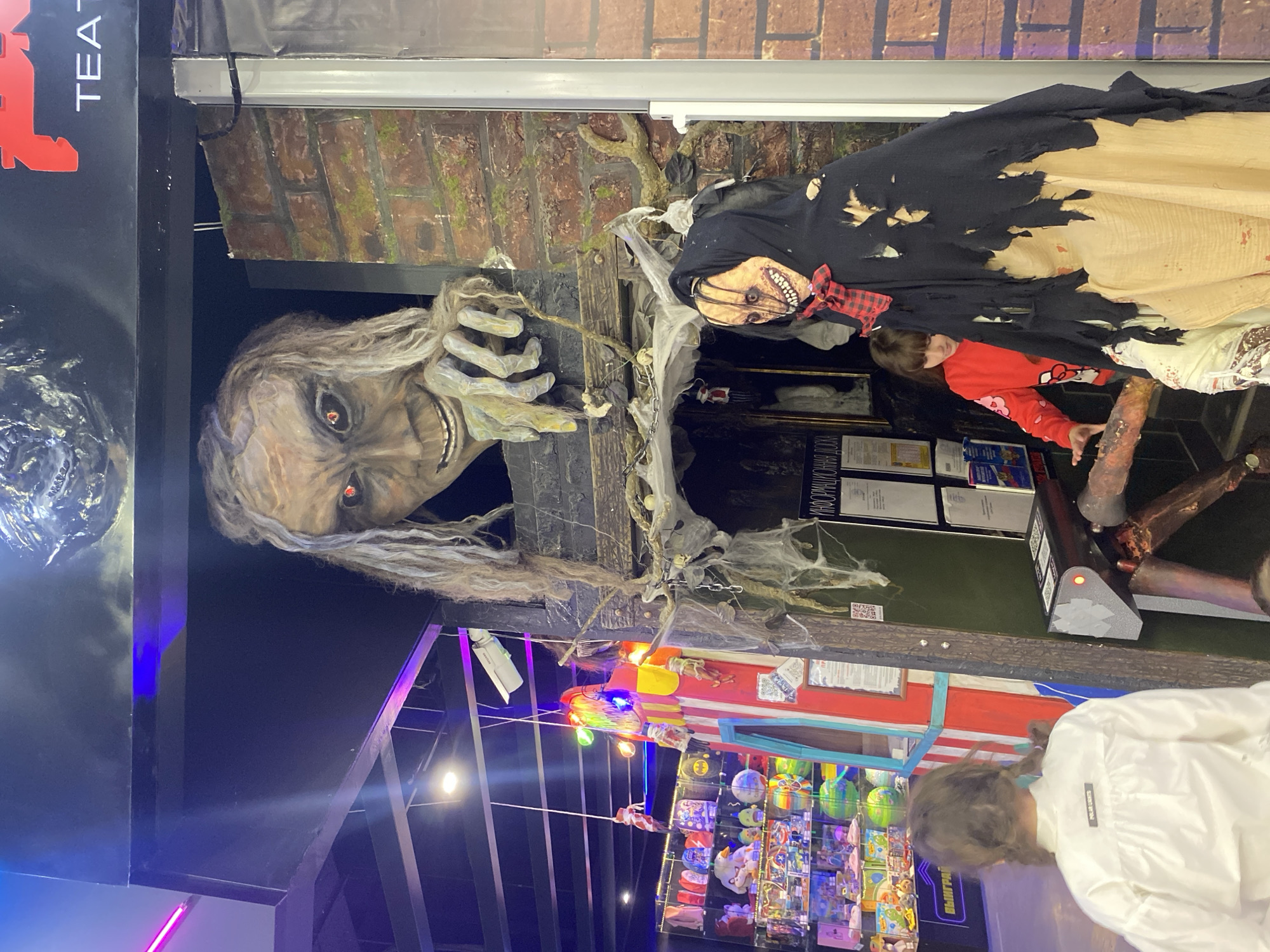 The image depicts a Halloween display in an indoor setting. The location is possibly in Yekaterinburg, Russia, as evidenced by the Cyrillic text "ИНФОРМАЦИЯ" on the sign. There is a monstrous, ghoulish figure with long, stringy hair and a wide, menacing grin. There is a second Halloween figure wearing a torn black shroud and a red plaid bow tie. A child in a red sweatshirt with a cartoon print is present, appearing to interact with the display. Shelves filled with colorful items, likely for sale, are visible below the horror display. One individual is wearing a white jacket. The background includes what appears to be a faux brick wall with some artificial vines. Overhead lighting and other structural elements are also visible.