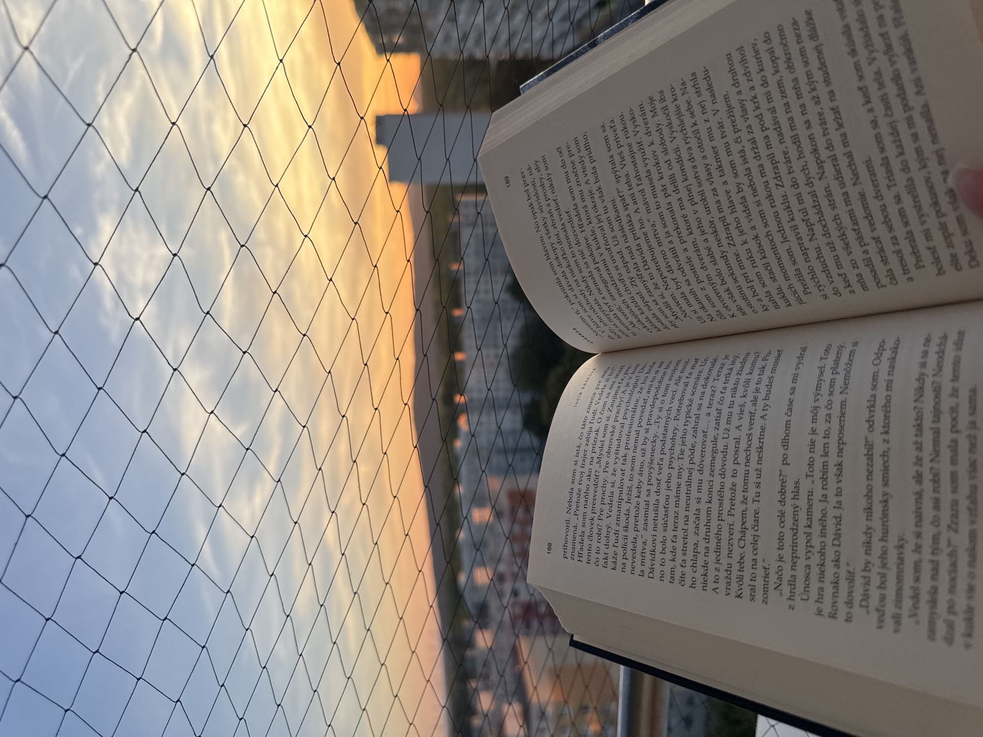 A close-up shot shows an open book placed on a ledge, with a protective net and a city skyline in the background. The sky displays a warm sunset gradient of blue and orange. The book's pages are open to text in Slovak, with page numbers 188 and 189 visible. The ledge is likely a balcony or windowsill, part of an apartment building in Bratislava, Slovakia, given the architectural style and urban setting. The composition suggests a moment of quiet contemplation, with the book serving as the central subject against the backdrop of the city at dusk.