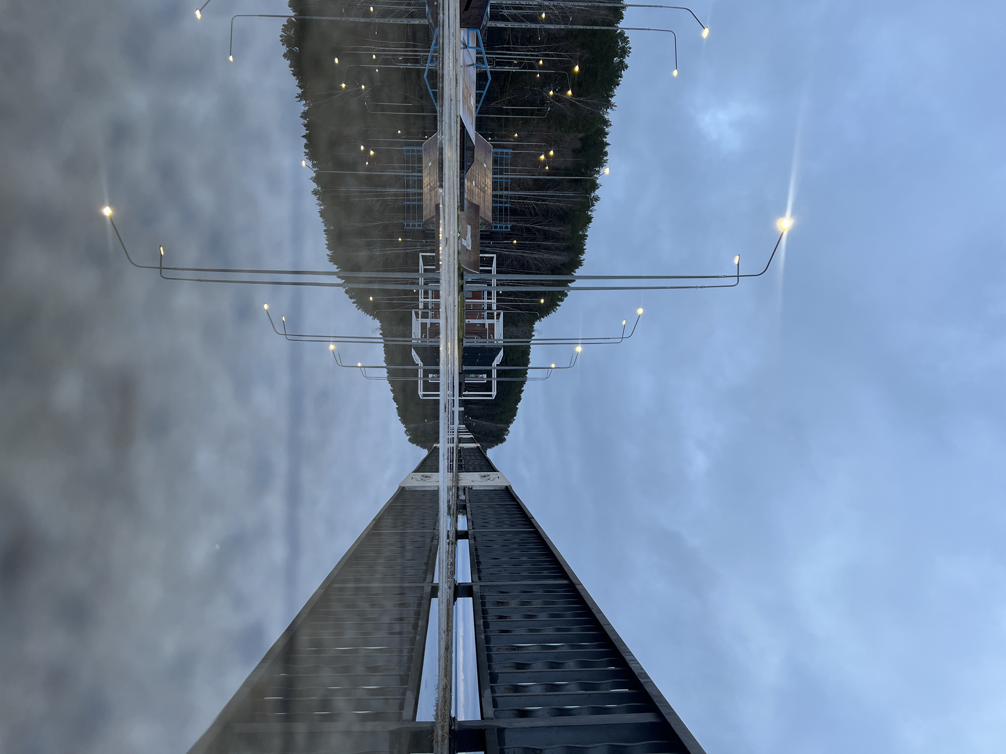 The image shows a view from the bottom of a ski jump in Chaikovsky, Russia. The ski jump's metal structure ascends towards the top, with a wooden walkway in the middle. Street lights line the sides of the ski jump. The view from the top of the ski jump is reflected in the water. Above the reflection is a cloudy sky.