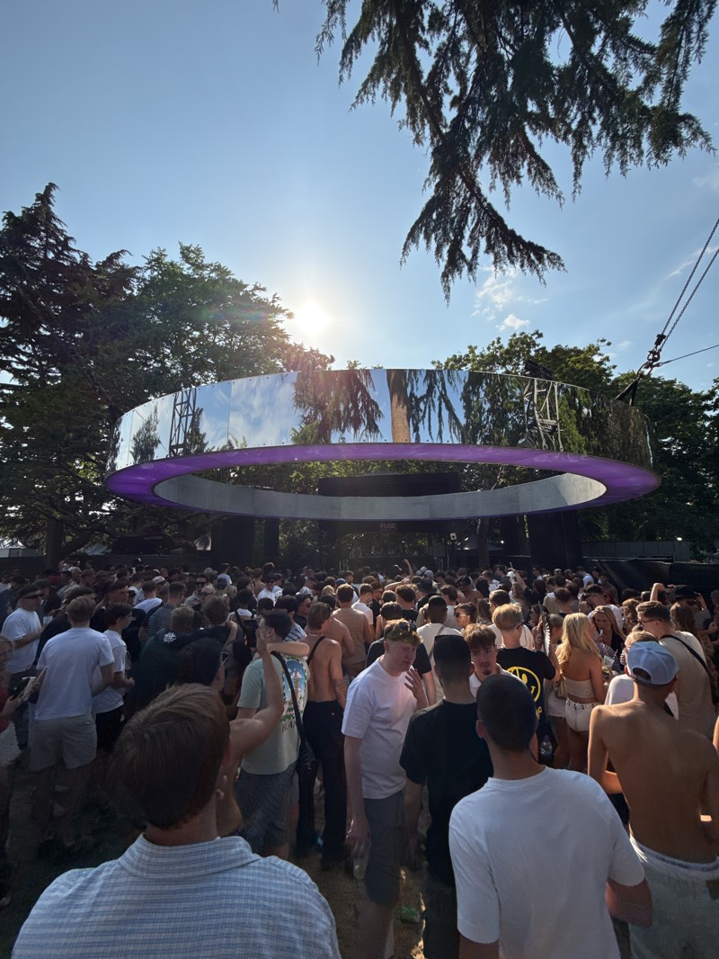 The image depicts a large crowd gathered at an outdoor event in Barking, United Kingdom. Suspended above the crowd is a large, reflective, circular structure with a purple light underneath. The crowd is composed of numerous people of varying ages and attire. Trees are visible in the background. The sky is clear, and the sun is shining brightly.