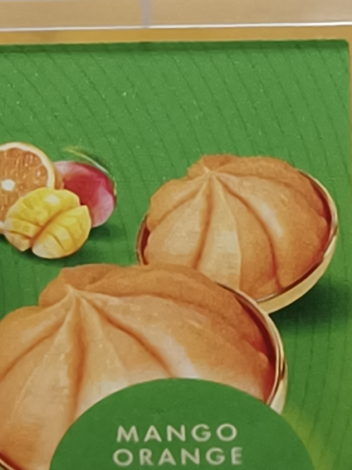The image shows a product package, likely for a food item or cosmetic. The background is green with a grooved texture, possibly simulating a leaf. In the upper left corner, there are cut segments of an orange and mango, along with a red radish. The main focus of the image is two dome-shaped objects with segmented surfaces and a light orange color. They are contained within small bowls with golden rims. A green circular label partially obscures the bottom object, with the text "MANGO ORANGE" visible. No people are present. No information is available to determine the location as Kryvyi Rih, Ukraine.