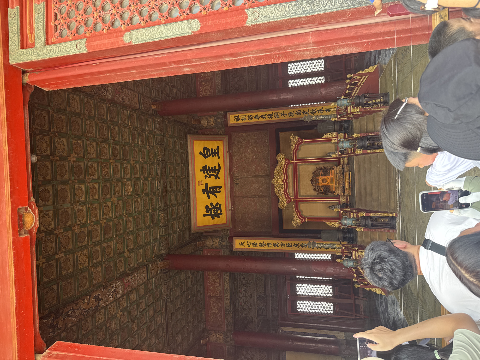 The image captures an interior view of a grand hall, likely part of the Palace Museum in Dongcheng District, China. Dominating the scene is an ornate golden throne situated on a raised platform, accessed by a set of red stairs with golden railings. The throne area is richly decorated, with intricate carvings and traditional Chinese architectural elements visible throughout the hall, including a heavily ornamented ceiling with repeated circular motifs and dragons.
Several people are present, primarily in the foreground, facing away from the camera towards the throne. They appear to be tourists, with some holding up smartphones to take photographs. One individual in the foreground is wearing a white t-shirt and has a black strap across their shoulder, suggesting a bag. The architectural setting is characterized by prominent red pillars and beams, painted red wooden doors with decorative lattice work, and large windows with similar lattice patterns. Two vertical yellow banners with Chinese calligraphy hang on either side of the throne area. The overall atmosphere suggests a historical and significant location, likely a place of imperial power or ceremony.