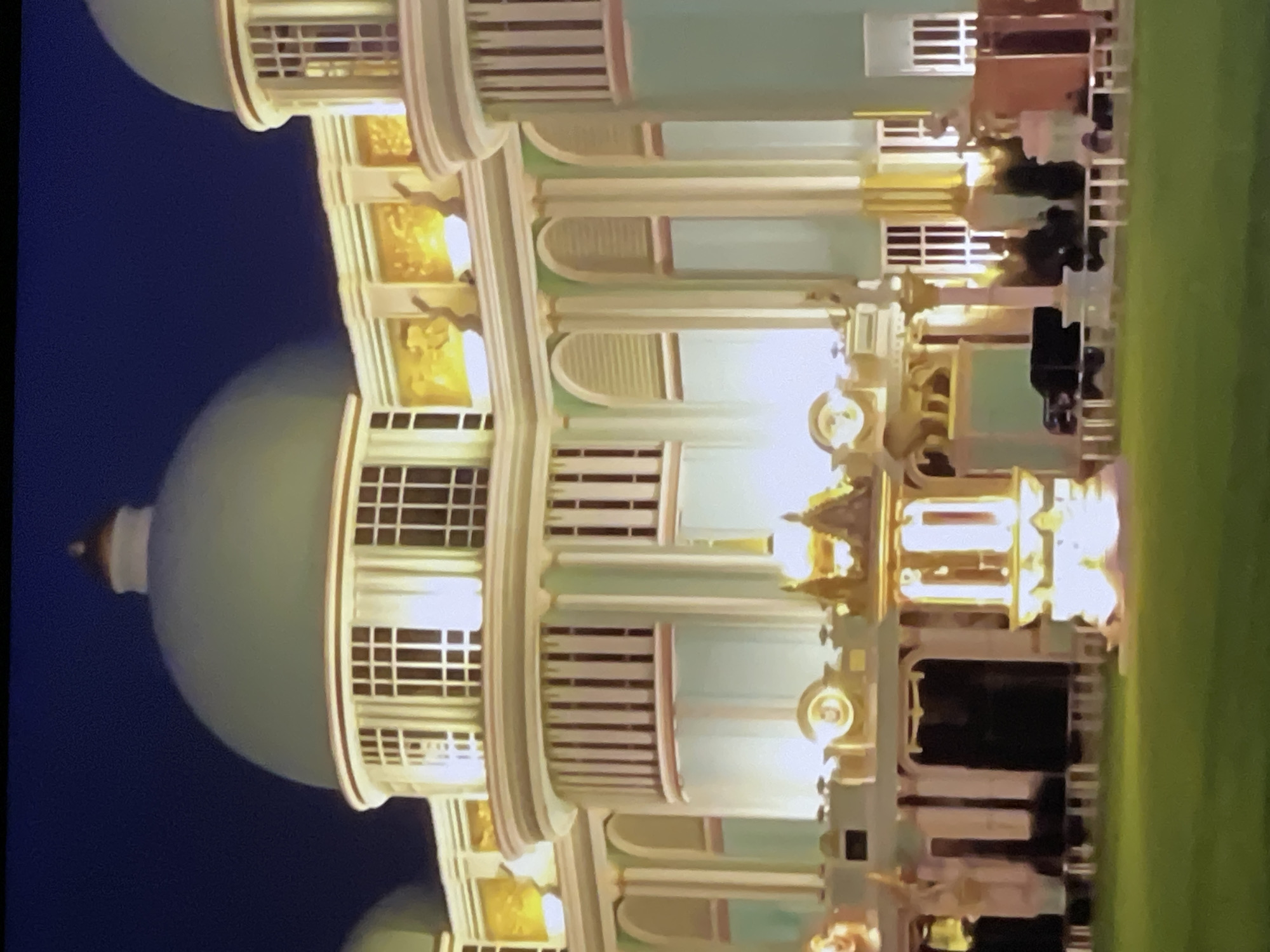 The image shows a close-up, slightly blurry, nighttime view of a grand building with multiple domes and ornate architectural features. The building appears to be lit up from within and with external spotlights, highlighting its pastel green and cream color scheme, with gold accents. The structure has a classical or baroque style, featuring arched windows, columns, and decorative reliefs. In the foreground, a well-maintained green lawn is visible, and in the middle ground, several figures and what appear to be vehicles are present, though their details are indistinct due to the image quality and lighting. One prominent element in the foreground is a brightly lit, ornate pavilion-like structure. The sky above is a deep, dark blue, indicating it is nighttime. This building is Sanctuary of Truth in Pattaya City, Thailand.