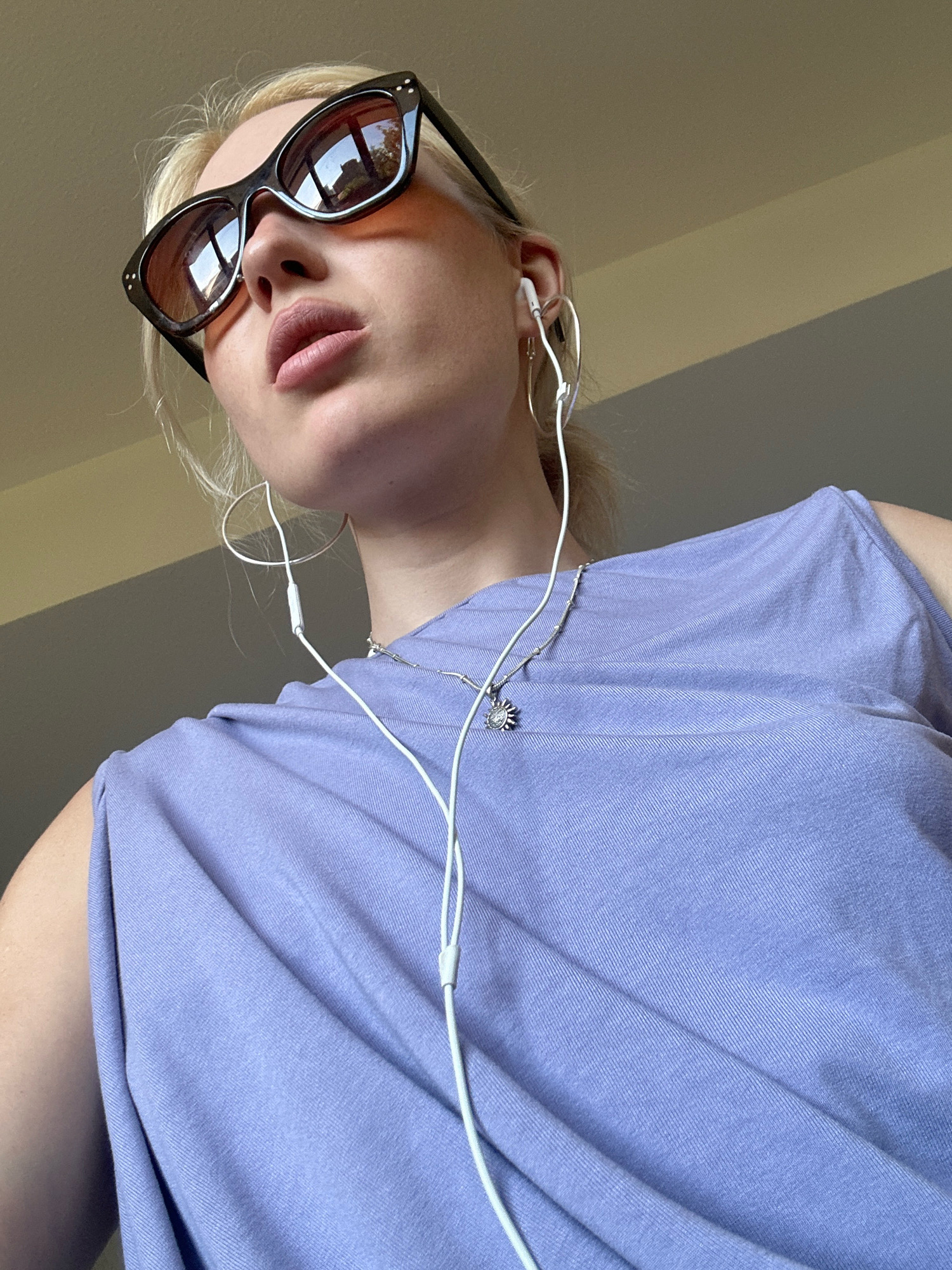 The image shows a close-up, low-angle shot of a young blonde woman wearing sunglasses and a light purple sleeveless top. She is also wearing white earbuds and a silver necklace with a small pendant. The background appears to be an indoor setting with a plain wall. The lighting suggests it might be daytime, possibly near a window. There is no discernible indication of the city of Tbilisi, Georgia, or any specific urban environment.