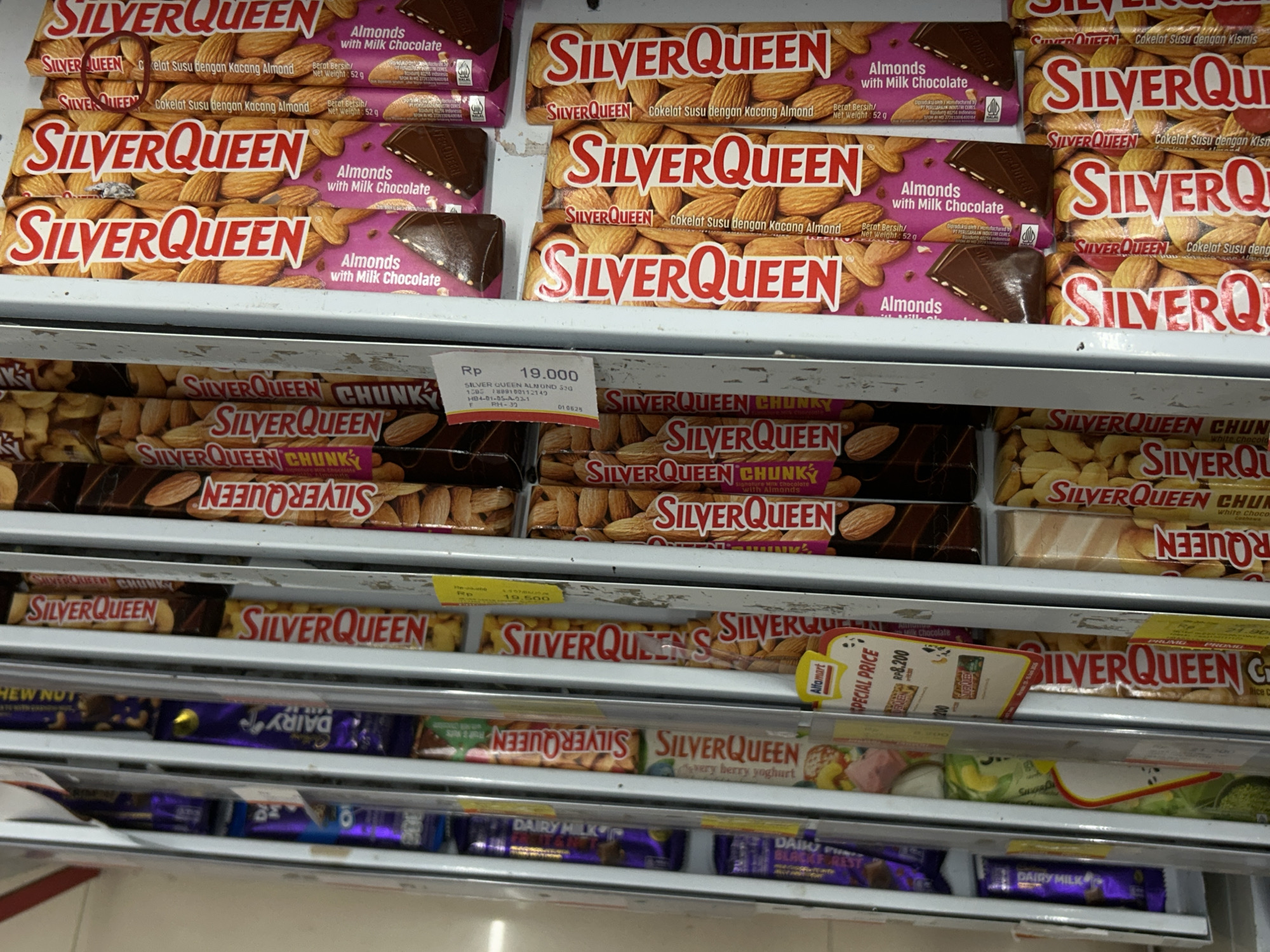 This image captures a close-up view of chocolate bars displayed on shelves in what appears to be a supermarket or convenience store in Payadua, Indonesia. The shelves are well-stocked with various brands and flavors of chocolate.
The predominant brand visible is "SilverQueen," with numerous bars featuring "Almonds with Milk Chocolate" or "Cokelat Susu dengan Kacang Almond." Many of these bars are labeled with "Berat Bersih / Net Weight: 52 g." Another variant, "SilverQueen CHUNK," is prominently displayed, including flavors like "Milk Chocolate with Almonds" and "White Chocolate." Other SilverQueen options include "Cokelat Susu dengan Kismis" (Milk Chocolate with Raisins), "very berry yoghurt," and "Rice Crispy" (labeled "SILVER QUEEN G").
Price tags are visible, indicating costs in Indonesian Rupiah (Rp). One SilverQueen bar is priced at "Rp 19.000," another at "Rp 19.500," and a "SPECIAL PRICE" tag from "Alfamart" shows an item at "Rp 8.200." Below the SilverQueen products, bars of "DAIRY MILK" chocolate are also visible, including a "DAIRY MILK BLACK FOREST" variant. The presence of Indonesian language on product packaging and the "Rp" currency symbol, along with the "Alfamart" logo, reinforce the location context of Indonesia. The scene suggests a typical retail environment, showcasing products available for purchase.