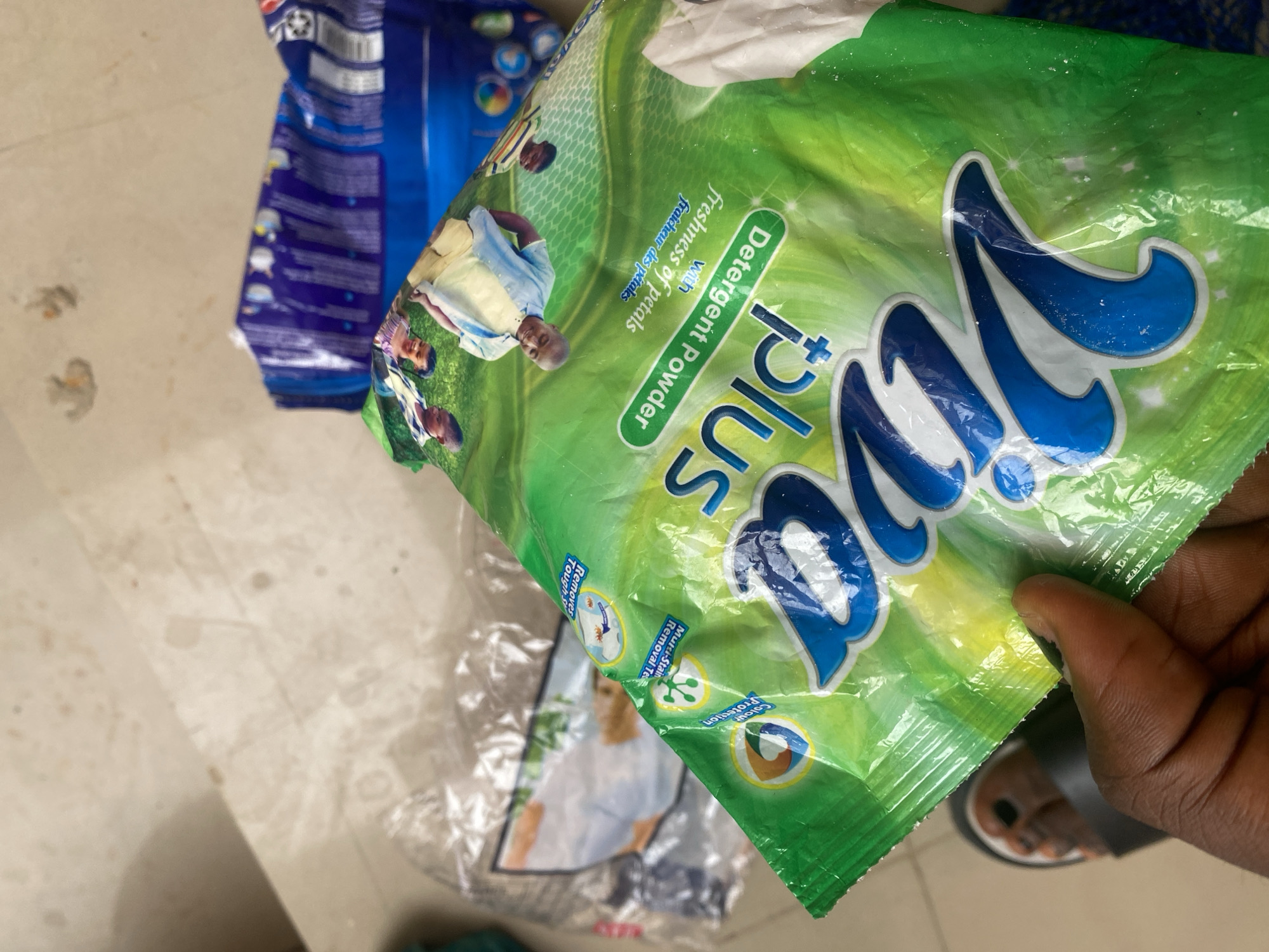 This is a close-up shot of a green and white packet of "Viva plus" detergent powder. The packet is slightly crinkled and appears to be held by a person's hand, with a foot wearing a sandal visible at the bottom right corner. The packet has large blue lettering spelling out "Viva plus". There are images of people on the packet, including what appears to be a family. Text on the packet indicates it is a "Detergent Powder" and mentions "freshness of petals" in both English and French ("fraîcheur des pétales"). There are also smaller icons and text highlighting features like "Colour Protection", "Multi-Stain Removal Tech", and "Removes Tough S". In the background, another blue detergent packet is partially visible. The setting appears to be indoors with a tiled floor. The lighting is bright, suggesting it is daytime. The overall impression is that of a product being displayed or handled.