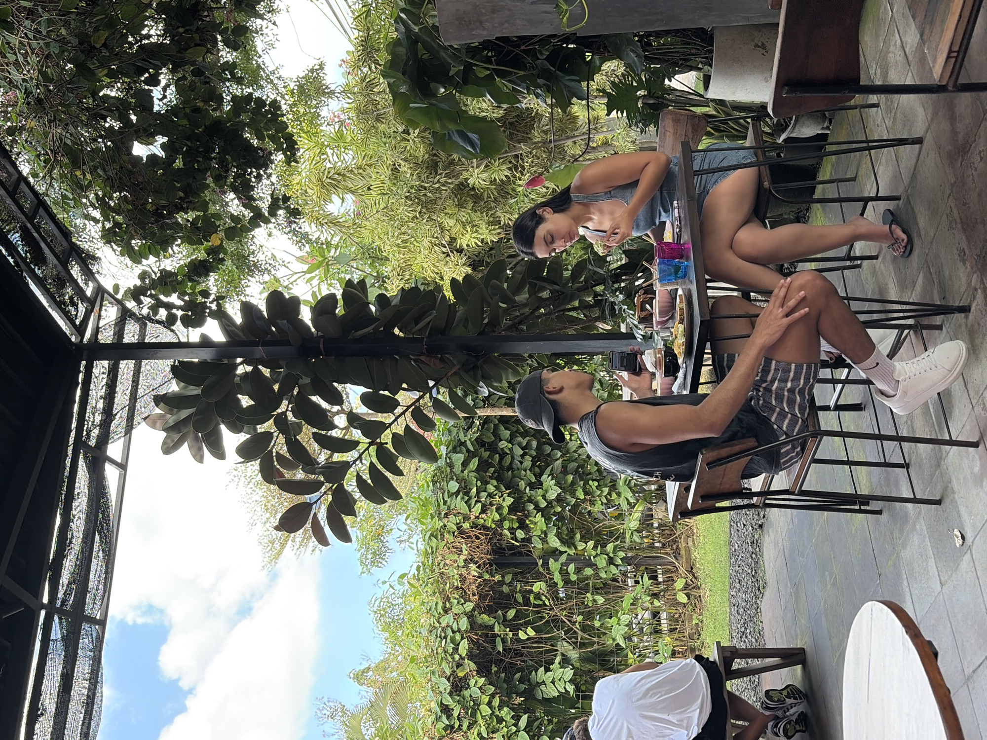 In North Kuta, Indonesia, two individuals, a man and a woman, are seated at a table in an outdoor cafe, engaged in a meal. The man, wearing a dark tank top, shorts, and a baseball cap, is looking down at his phone. The woman, dressed in a denim overall dress, is eating. On the table between them are plates of food, chopsticks, and colored glasses. They are positioned on a patio with a tiled floor, surrounded by lush greenery and trees. A portion of another person, wearing a white shirt and dark shorts, is visible in the background, seated on a wooden bench. The scene is captured under a canopy, suggesting a covered outdoor seating area.