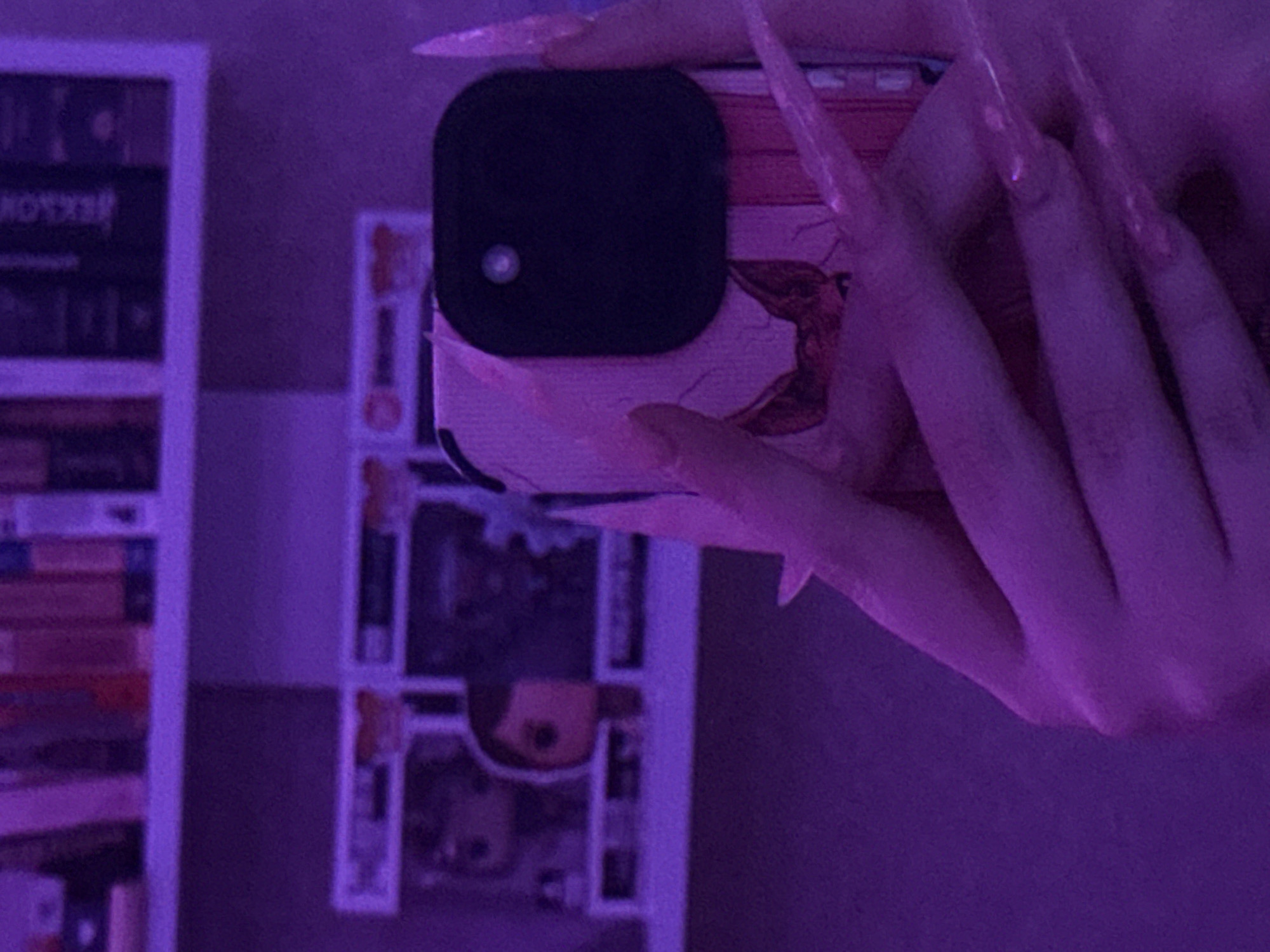 The image is a close-up, low-angle shot of a person's hands holding a phone. The phone has a black camera module and a pink case with a graphic of a cat's face. The person has very long, pointed fingernails, which are translucent and appear to be adorned with glitter or similar material. The overall lighting of the scene is a deep purple hue, suggesting it might be illuminated by colored lights or is a nighttime shot.
In the background, there are shelves filled with books and what appear to be collectible figurines, possibly Funko Pops, inside clear plastic boxes. The setting is indoors, with the purple lighting creating a somewhat atmospheric or stylized effect. There are no visible clues to suggest the location in Pudomyagi, Russia.