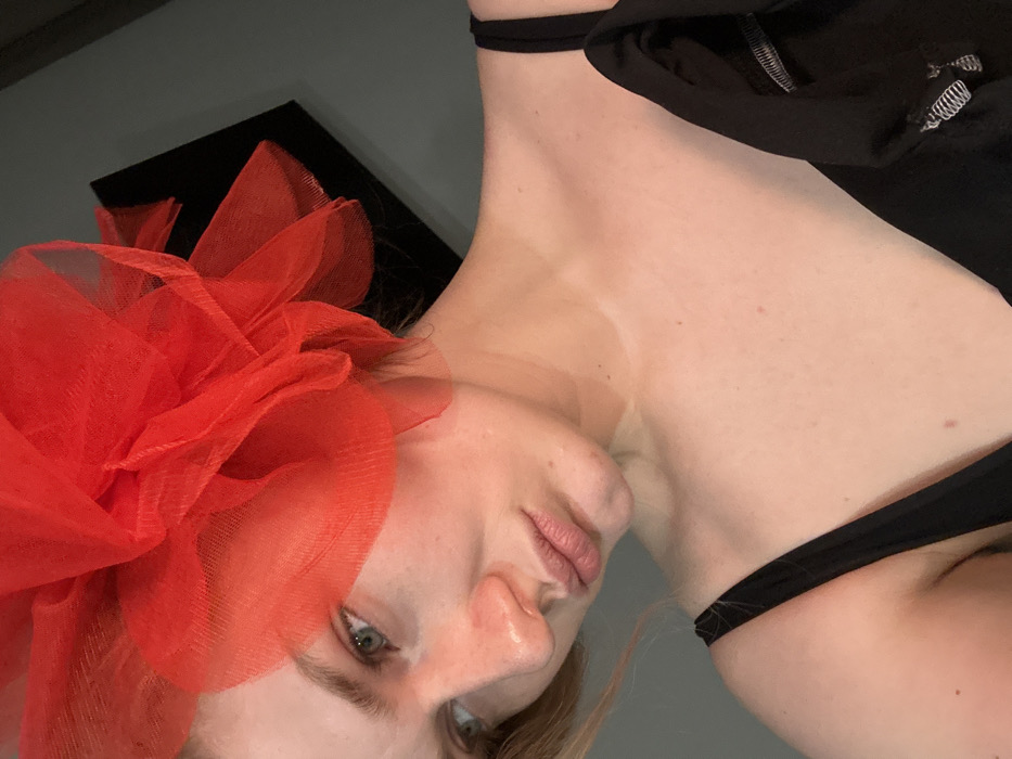 The image displays a close-up, slightly upward-angled indoor view of a fair-skinned adult female, located in Kazan, Russia. She has light-colored hair, partially visible, and green eyes, with her gaze directed towards the upper left. A large, voluminous hair adornment made of layered sheer red fabric is positioned on the right side of her head. She wears a black top with thin straps, partially obscured by a black outer garment draped over her left shoulder, which features a visible silver-toned zipper pull. The background consists of a plain, dark gray wall, against which a tilted black rectangular object is visible in the upper right.