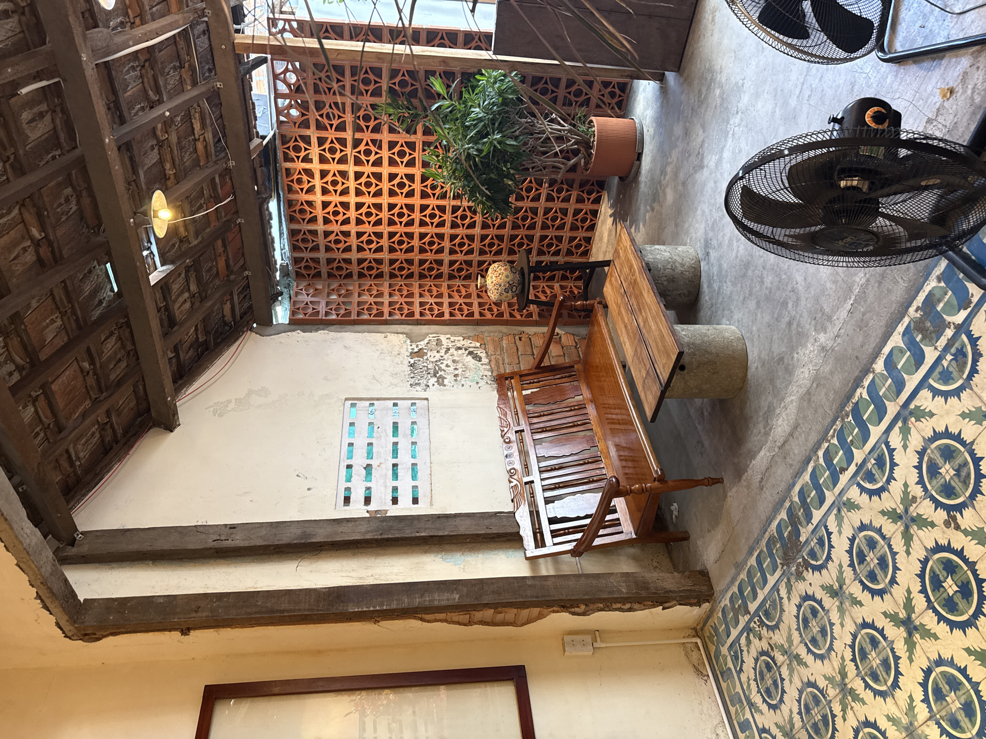 The image depicts an outdoor seating area of a cafe in Bac Nha Trang Ward, Vietnam. A wooden bench and coffee table are situated on a concrete floor with a patterned tile border. To the right, a large potted plant stands in front of a decorative terracotta screen. In the foreground, two black electric fans are visible. The background shows exposed brick walls and a rustic wooden roof structure with a hanging light fixture.