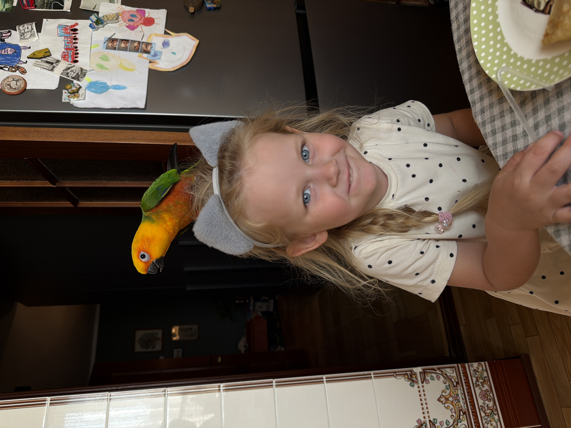 The scene is reportedly located in Khimki, Russia. A young blonde girl with blue eyes, wearing a cream-colored short-sleeved top with black polka dots and a grey cat-ear headband, smiles directly at the viewer. A colorful parrot, identified as a Sun Conure with orange, yellow, and green plumage, is perched on her head, positioned behind the cat ears and facing left. The girl's left hand rests on a grey and white checkered tablecloth. To her right, a green plate with white polka dots holds a piece of food, possibly a crepe with a dark topping, alongside a clear plastic straw. The background on the left features a vertical structure with white tiles adorned with intricate floral patterns in brown, pink, and green, adjacent to a dark wooden doorway opening into a dimly lit room. This room contains dark walls, a dark floor, and indistinct furniture and framed pictures. On the right, a dark refrigerator is covered with numerous children's drawings, papers, and small photographs attached by magnets.