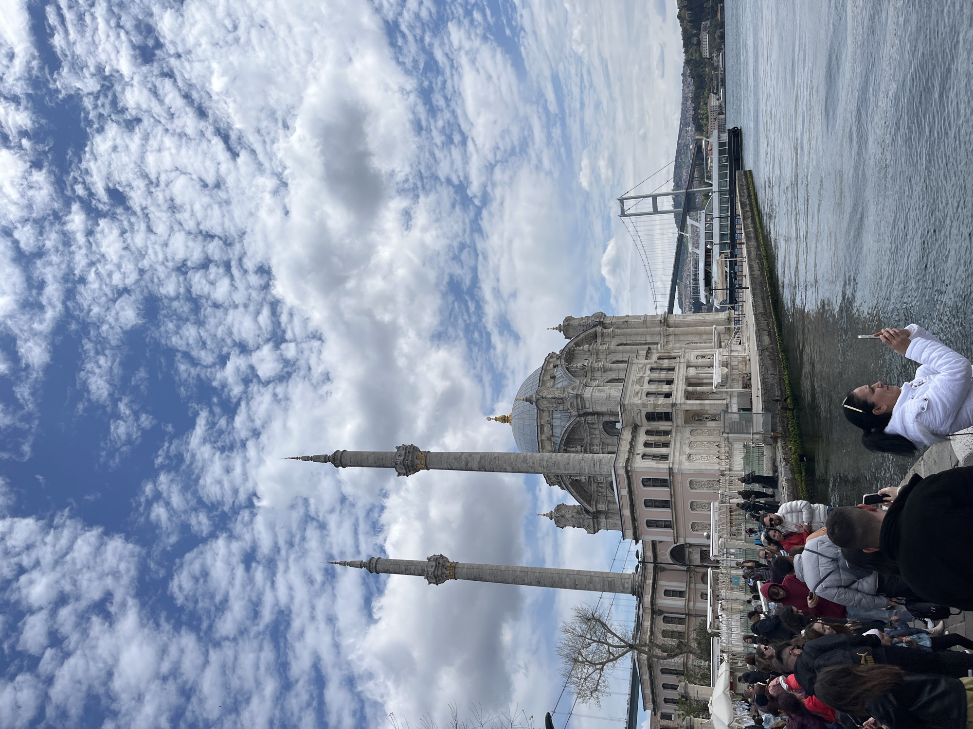 The image captures the Ortaköy Mosque in Beşiktaş, Turkey, under a cloudy sky. The mosque, with its two minarets, is positioned beside the Bosphorus Strait, with the Bosphorus Bridge visible in the background. A crowd of people is gathered near the mosque along the waterfront. Some are taking pictures.