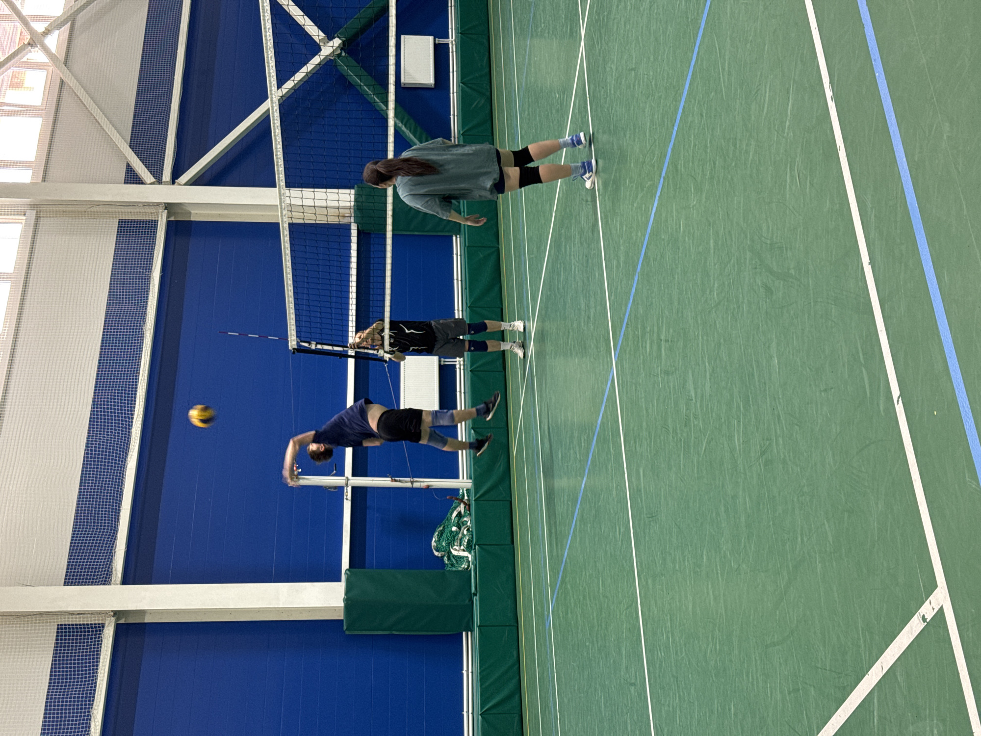 The image depicts an indoor volleyball court with three individuals playing. A male athlete in a dark blue shirt and black shorts is in mid-air, jumping to hit a yellow volleyball that is positioned above and to his right. He is wearing knee pads. On the other side of the net, a male athlete in a black t-shirt and grey shorts is standing, facing the net. He is also wearing knee pads. A female athlete in a teal t-shirt and dark shorts is standing further back on the same side of the net as the male athlete in black, also facing the net. She is wearing knee pads and has her hair in a ponytail. The court is green with white and blue lines marking boundaries. The background consists of a bright blue wall and a white ceiling with exposed beams and netting. The environment appears to be a sports complex or gymnasium. There is no discernible information in the image to indicate its location within the city of Saint Petersburg, Russia.