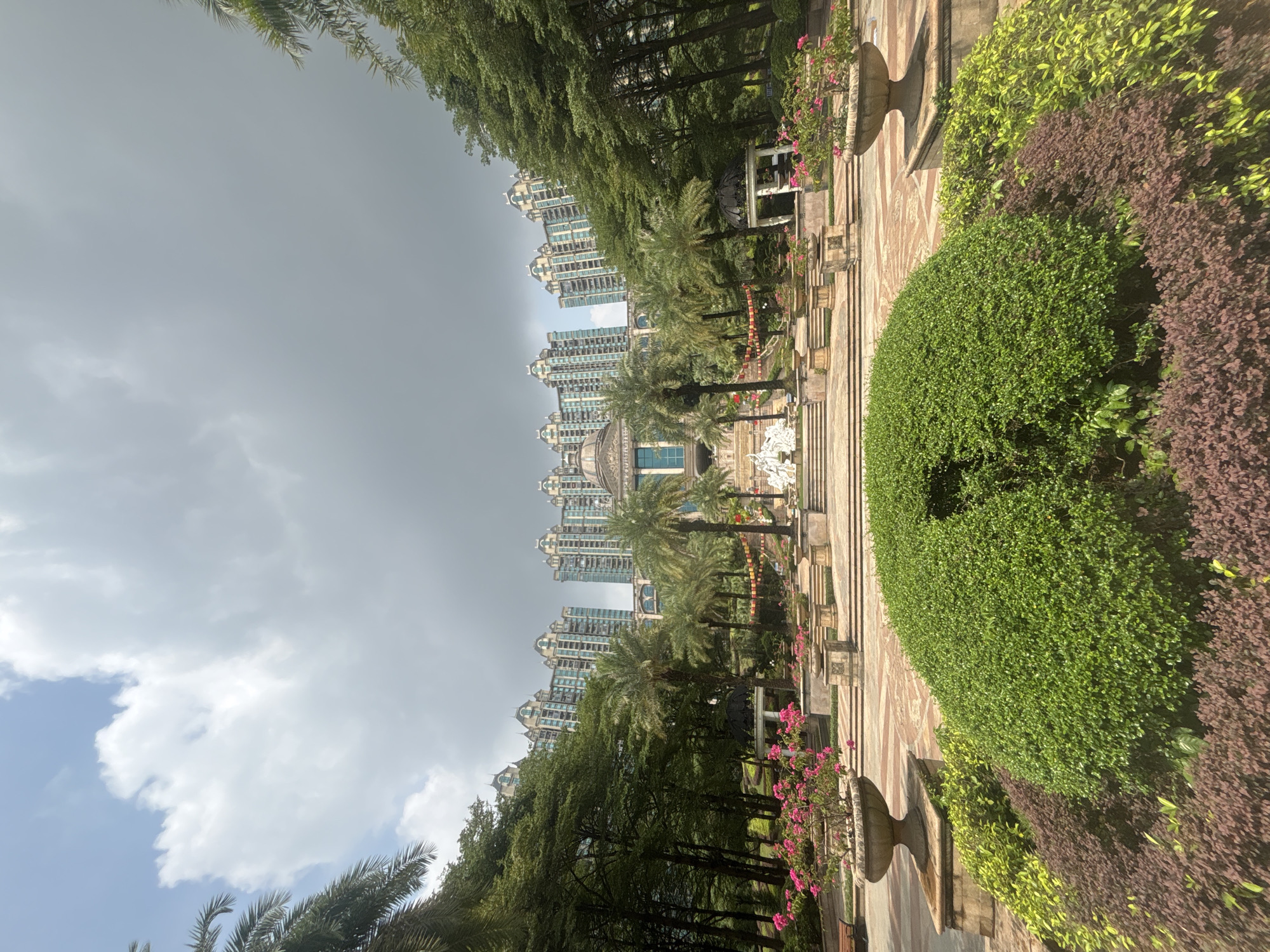 The image shows a wide-angle view of an opulent courtyard or garden area in the Nanhai District of China. In the background, a row of tall, ornate apartment buildings with distinctive domed roofs and light blue facades dominate the skyline. These buildings are set against a cloudy sky.
In the midground, a formal garden features a central fountain with white sculptures, flanked by palm trees and lush green foliage. A grand staircase leads up to a building with classical architectural elements and a dome. Red lanterns are visible hanging along the staircase, suggesting a festive or ceremonial atmosphere. White gazebos are situated on either side of the courtyard.
The foreground is occupied by meticulously sculpted hedges and bushes, with a large, rounded green bush taking center stage. To the left and right of this central bush are planters filled with vibrant pink flowers. Stone urns and tiered planters are strategically placed throughout the garden. The pathways are paved with a patterned stone. The overall impression is one of a well-maintained, luxurious residential or public space. No people are visible in the image.