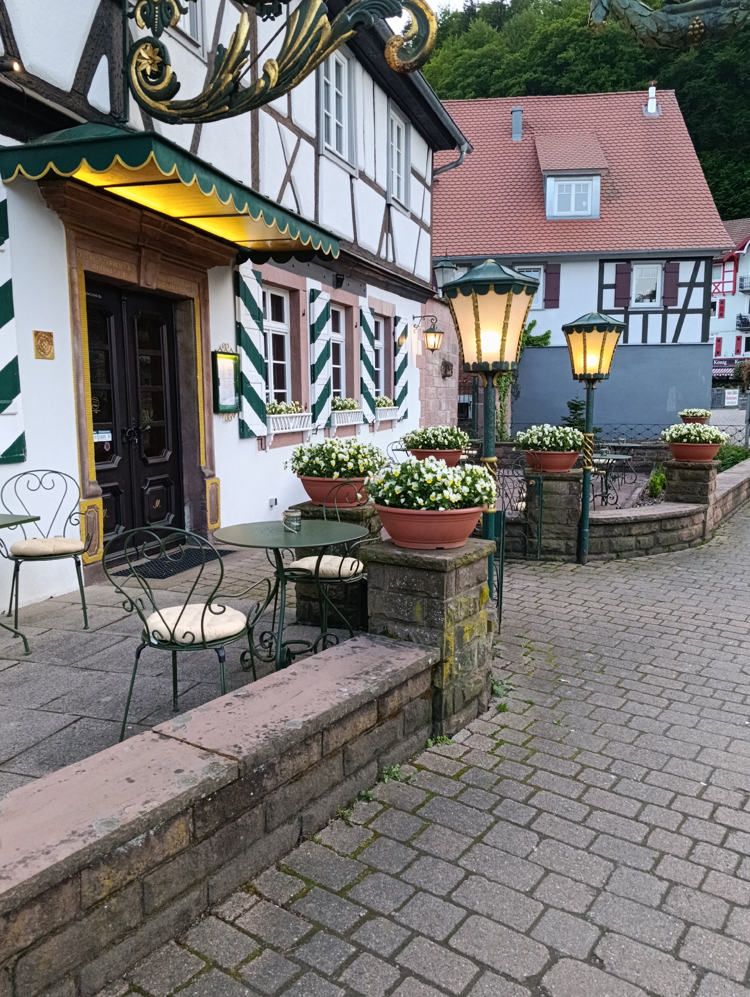 This image shows the exterior of a building with half-timbered architecture, characteristic of many European towns. The building has white walls with exposed dark wooden beams, and windows with green and white striped shutters. In front of the building, there is a small outdoor seating area with green metal tables and chairs. The seats have beige cushions. Above the entrance, there's a green awning with decorative gold trim and a matching gold design above.
To the right of the main building, another building is visible with a red tiled roof and dormer windows. A row of decorative lampposts with flower pots stands along a low stone wall leading to the seating area. The lampposts are lit. The ground is paved with cobblestones. There is a hillside covered with trees in the background. The city is not explicitly named, so it is labeled as "Unknown, Unknown."