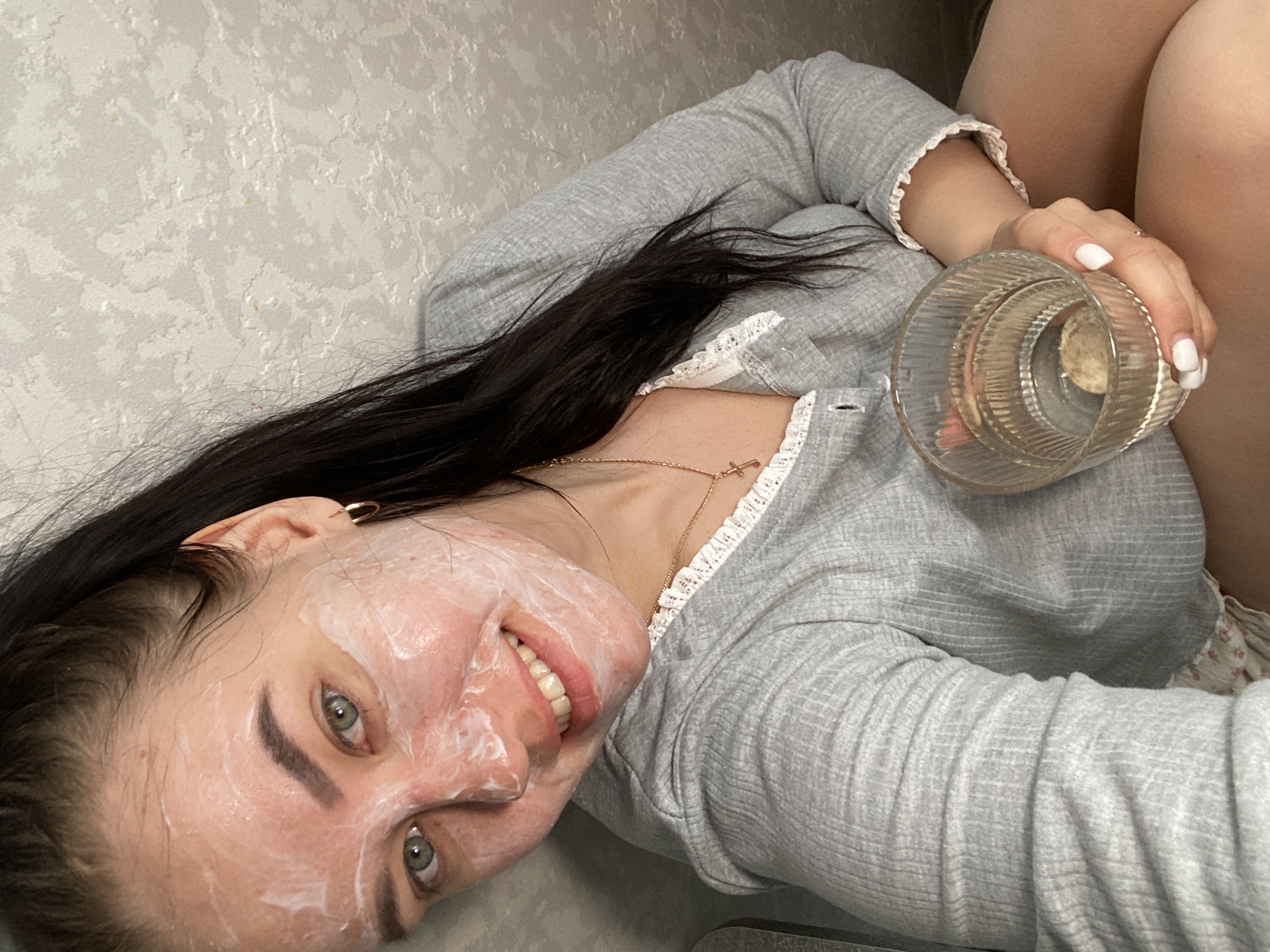 A woman is shown at close range in a low-angle, tilted, indoor shot. Her face is covered with a white substance, presumably a cosmetic face mask, but she is smiling. She has green eyes, dark hair, and fair skin. She wears a gray long-sleeved top with white lace trim. In her right hand, she holds a clear glass filled with water. She also wears a gold necklace. The background consists of a textured gray wall. The location is Chaikovsky, Russia.