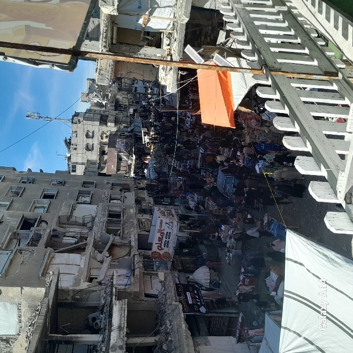 This image depicts a densely populated market street in Khan Yunis, Palestinian Territory, flanked by multi-story residential and commercial buildings exhibiting severe structural damage. Many structures display collapsed facades, exposed interiors, and broken windows, with rubble and debris visible on ledges and the street. The narrow street below is crowded with numerous individuals, primarily civilians, moving amidst makeshift market stalls. These stalls feature merchandise displayed on ground tarps and tables, often covered by white and orange tarpaulins providing shade. A black sedan is positioned within the crowded thoroughfare. Visible signage includes Arabic script, notably "الرملي" and "شركة أعمال العمري," with a UNICEF logo identifiable on a white tarp covering a section of a damaged building on the right. A cellular antenna tower is perched atop a distant building against a clear blue sky. The perspective is elevated, with a white wooden railing and a striped white tarp partially visible in the foreground.