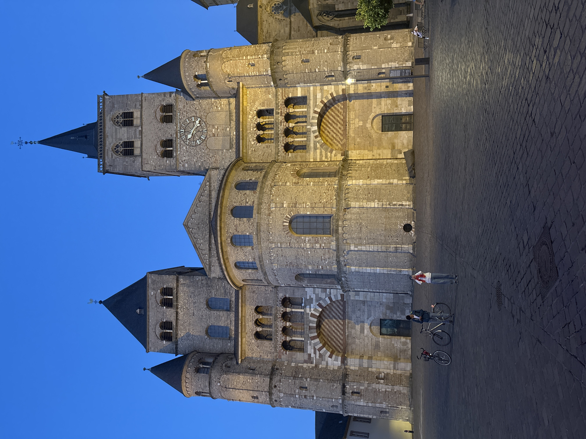 The image depicts the Trier Cathedral (Trierer Dom) in Trier, Germany, at dusk. The cathedral's Romanesque architecture is prominent, with its massive stone structure, twin towers, and arched windows illuminated by artificial lighting. In the foreground, a cobblestone plaza is visible. Two bicycles are parked near the center, and two individuals are present: one is standing near the bicycles, looking at their phone, and another person is standing further to the right, holding up their phone, possibly taking a picture or recording. The sky is a deep blue, indicating the twilight hours. Other architectural elements of the cathedral, including a clock face on one of the towers and smaller turrets, are also visible. The scene conveys a sense of quiet evening in a historic European city.
